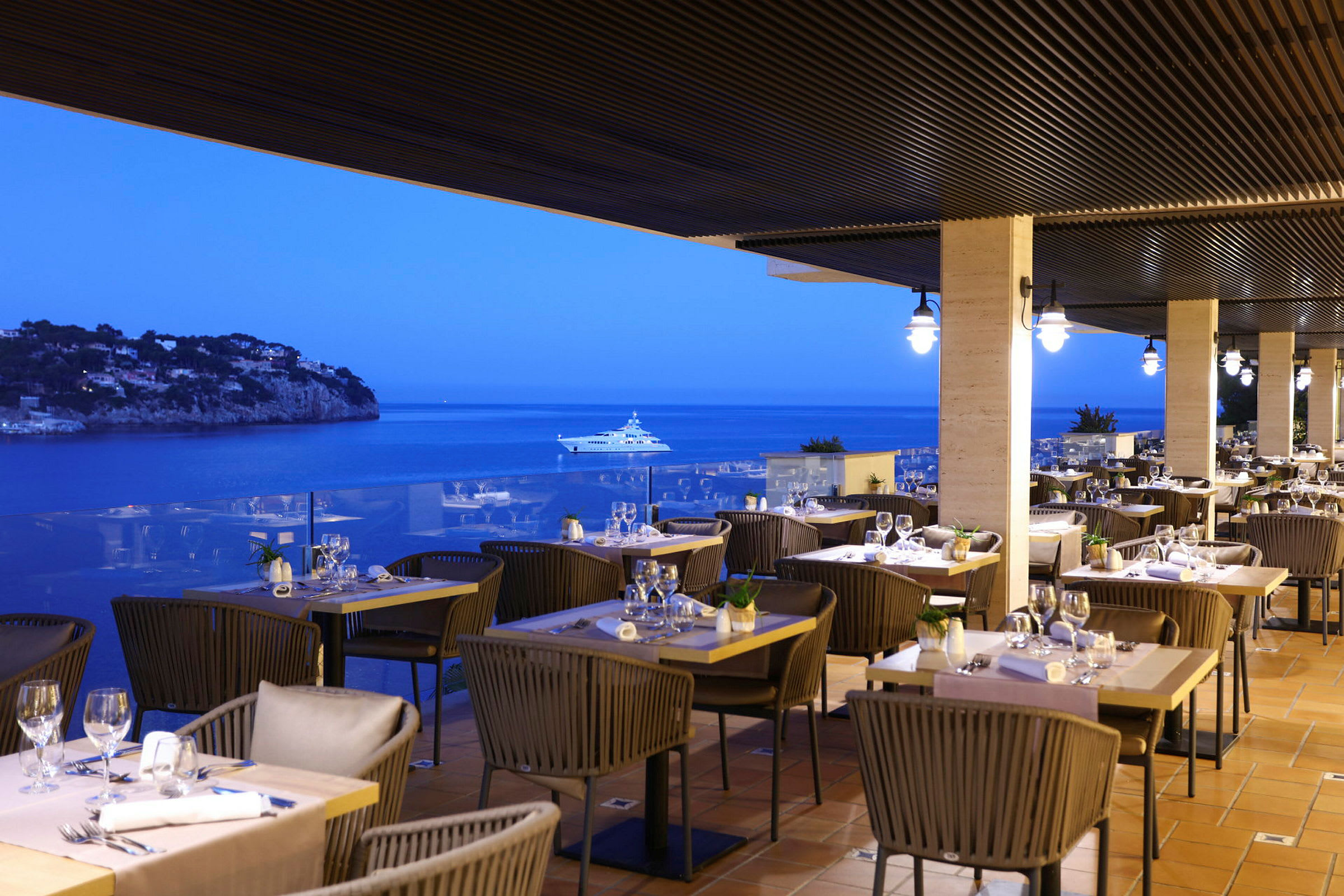 En restaurang med utomhusservering på en terrass med utsikt över havet på kvällen. Flera bord är uppdukade med stolar och glas. (AI-genererad text)