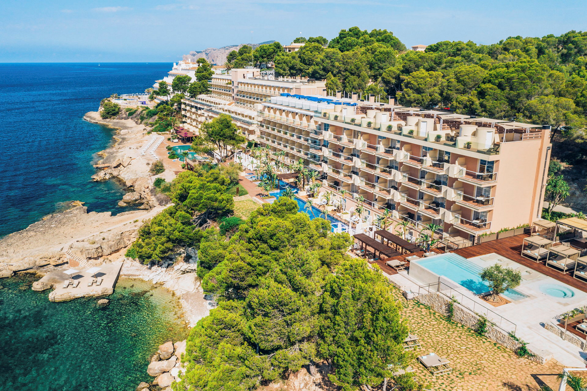 Fyrvåningshotell vid kusten med en swimmingpool framför och omgivande grönska. (AI-genererad text)
