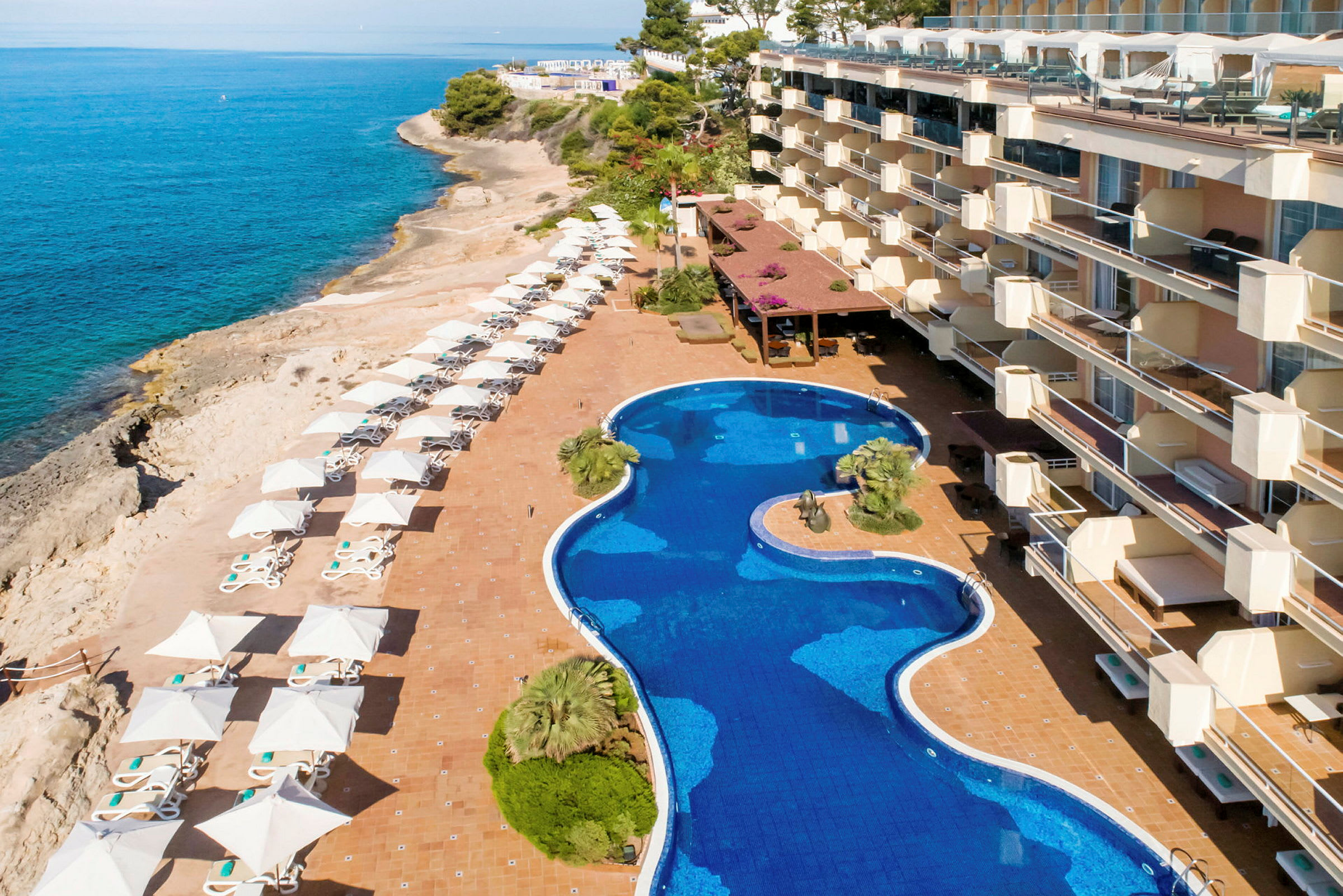 Bild av en hotellpool omgiven av solstolar och parasoller med utsikt över klippor och hav. (AI-genererad text)