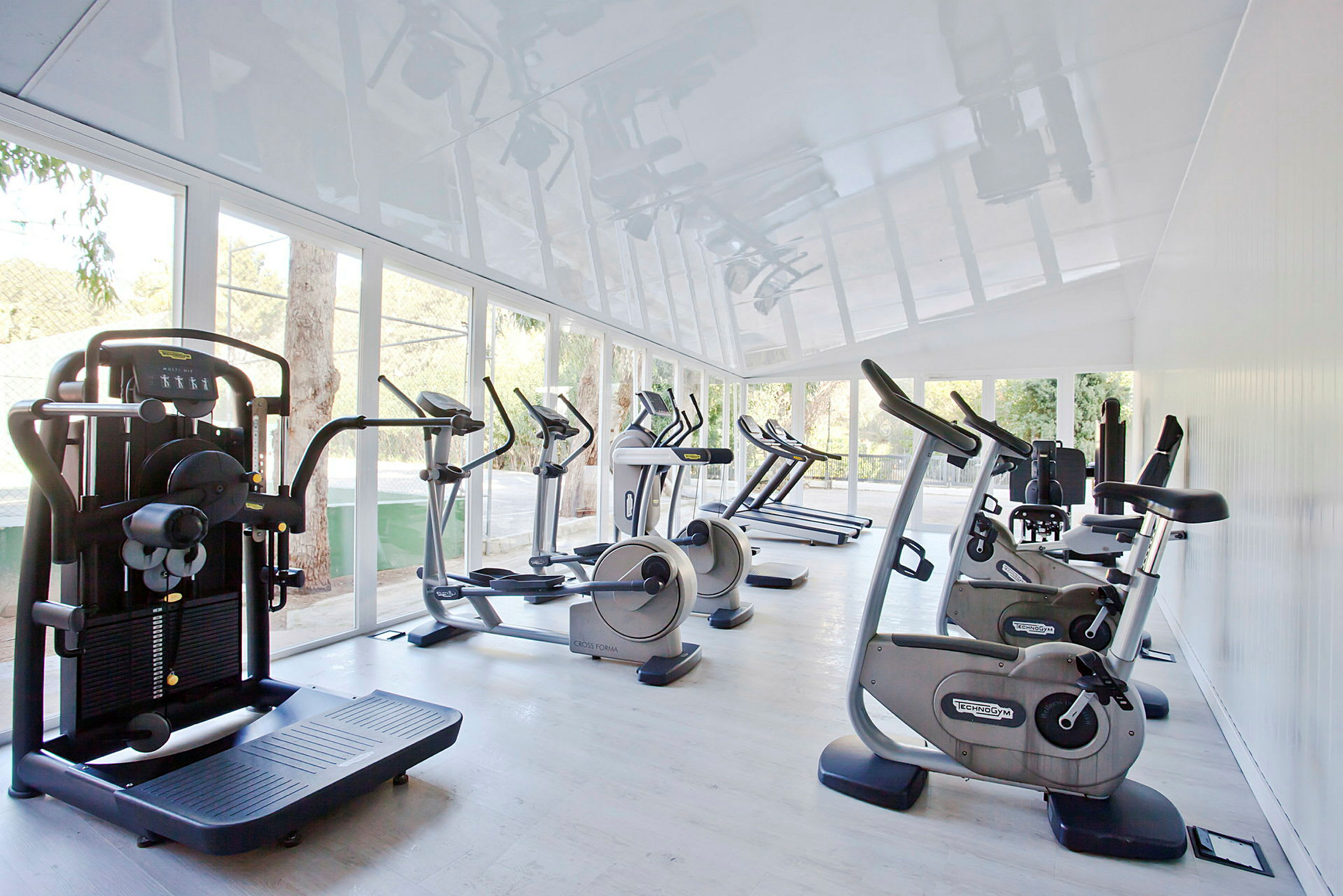 Ett gym med olika träningsmaskiner som motionscyklar och löpband placerade i ett ljust rum med stora fönster som släpper in naturligt ljus. (AI-genererad text)