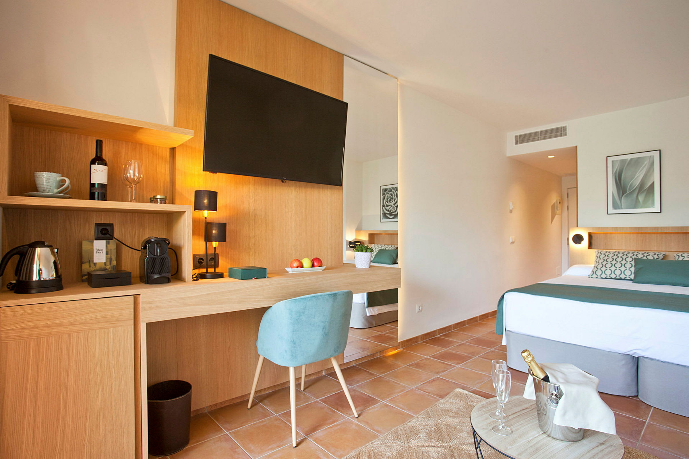 Ett modernt hotellrum med en dubbelsäng, en skrivbordsyta med stol, en väggmonterad TV och inkluderad kaffemaskin. Dekoration inkluderar ett fotografi och en vinflaska, samt en fylld champagnekyl på ett litet bord. (AI-genererad text)