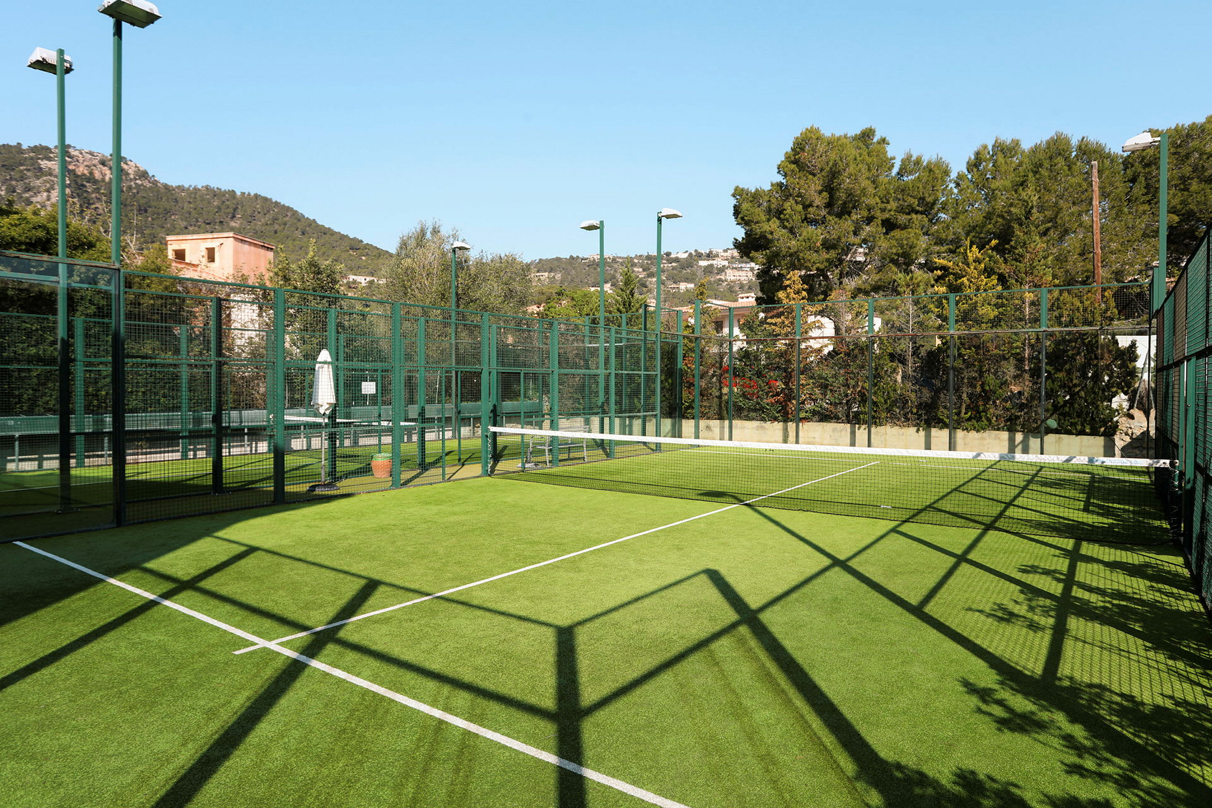 En padelbana utomhus med grön konstgräsbeläggning, omgiven av skyddsnät och omgiven av träd och byggnader. (AI-genererad text)