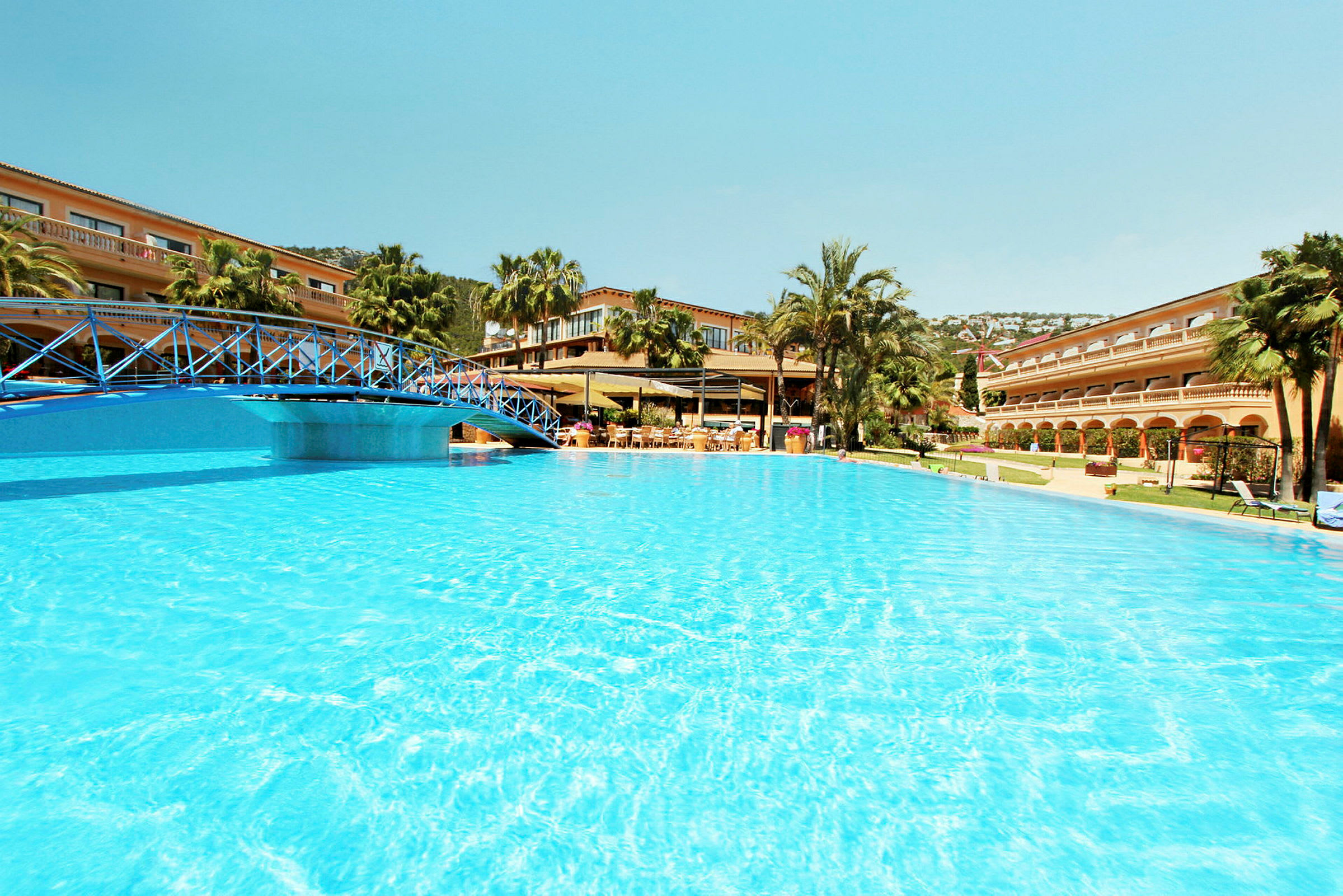 Bild som visar en pool vid hotellkomplex med en blå bro över vattnet och grästäckta omgivningar med palmer. (AI-genererad text)