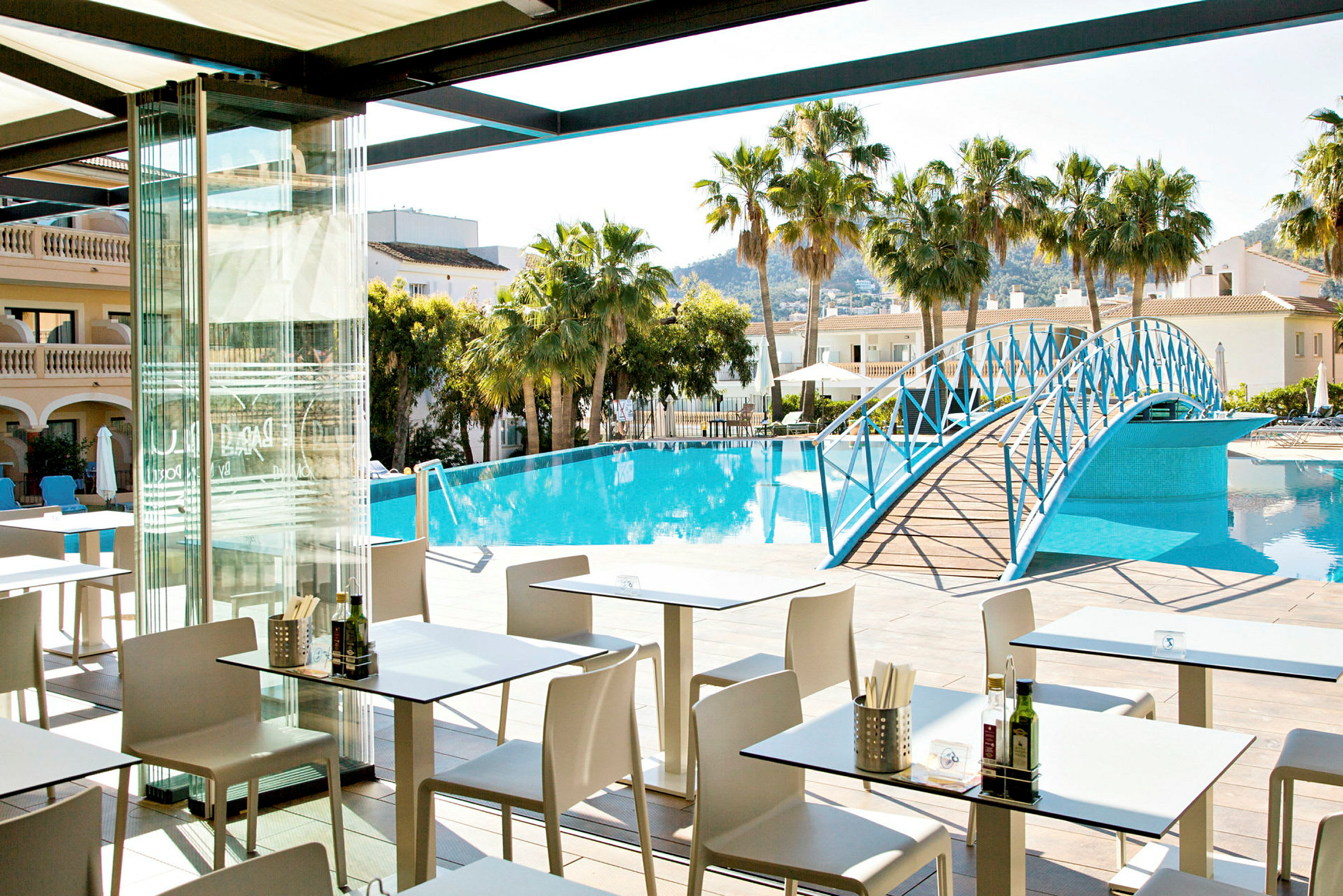 Ett restaurangområde med uteservering vid en pool omgiven av palmer och byggnader. En blå gångbro sträcker sig över poolen. (AI-genererad text)