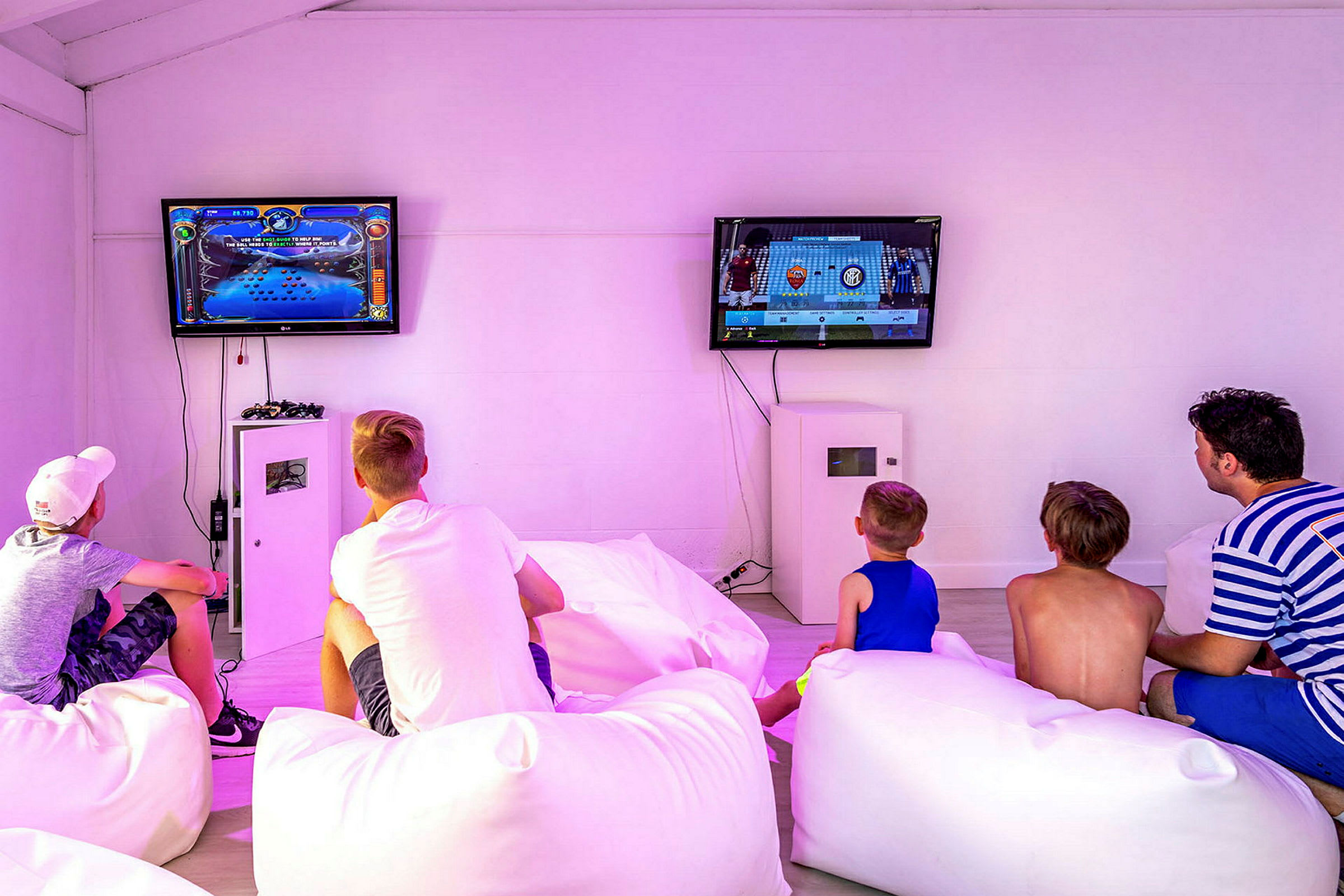 Personer sitter på vita kuddar och tittar på tv-spel på två skärmar. (AI-genererad text)