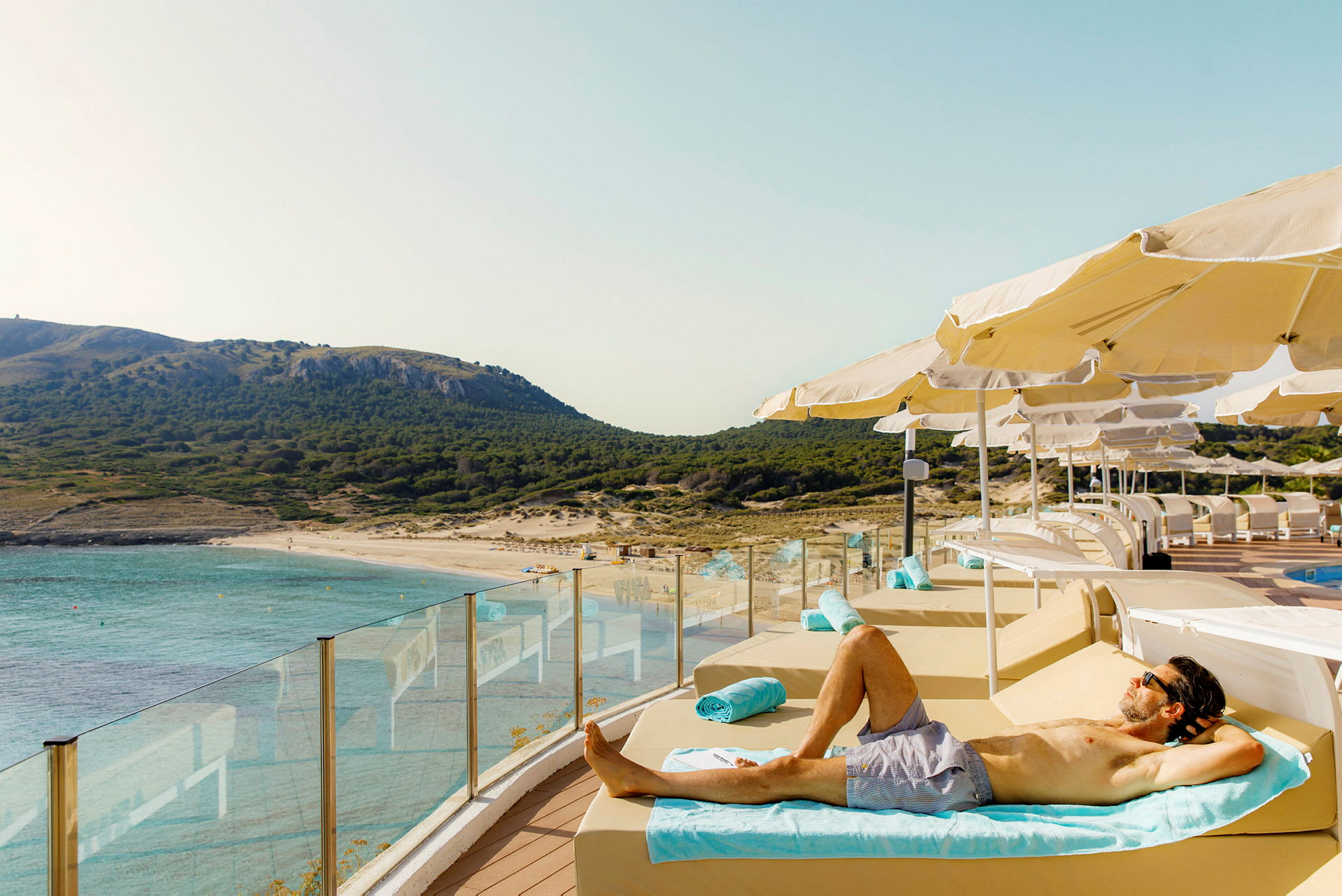 En person ligger på en solstol vid en pool med utsikt över en strand och bergig natur. (AI-genererad text)