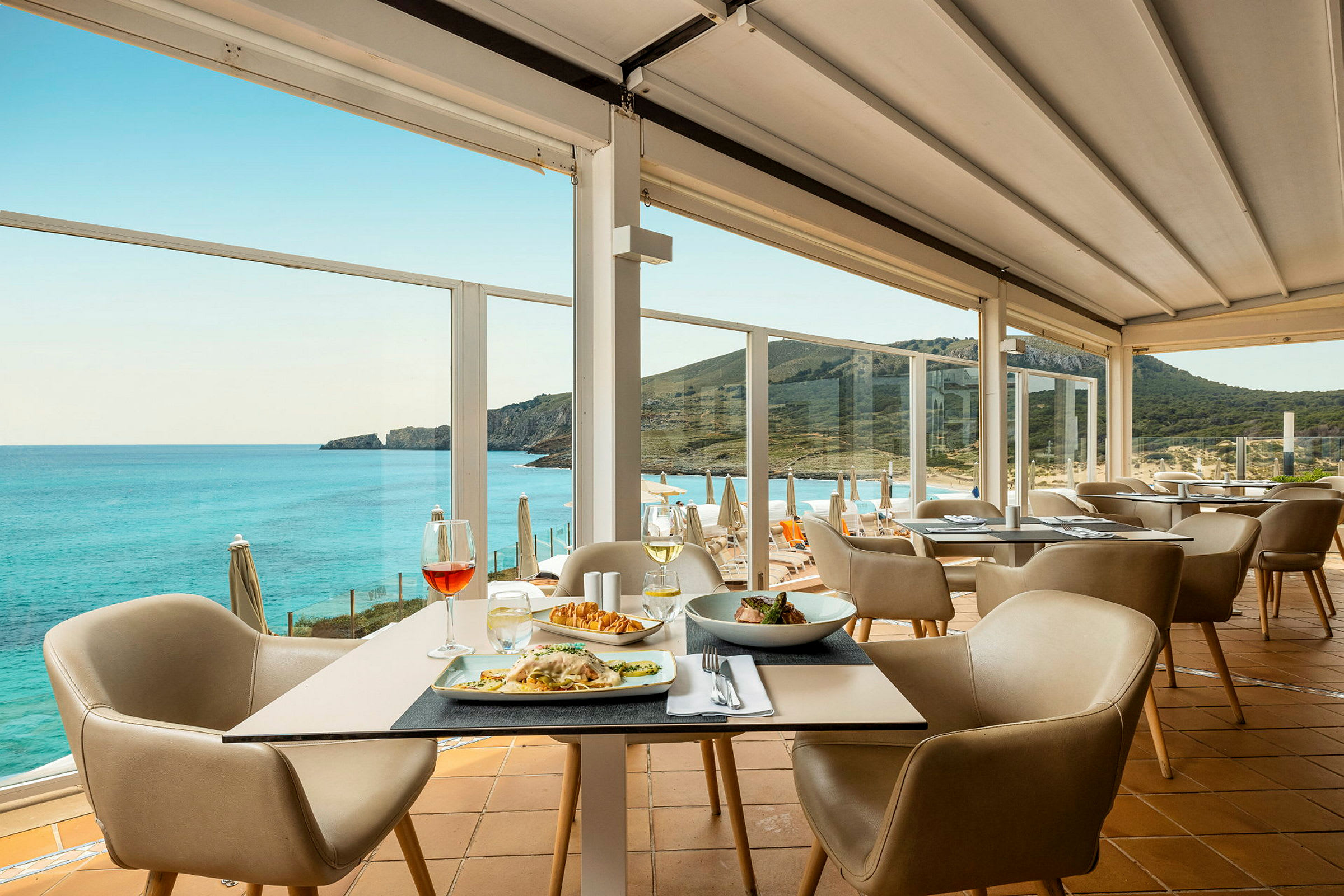 En restaurang med bord med mat och stolar, och en panoramautsikt över havet och en bergig kustlinje. (AI-genererad text)