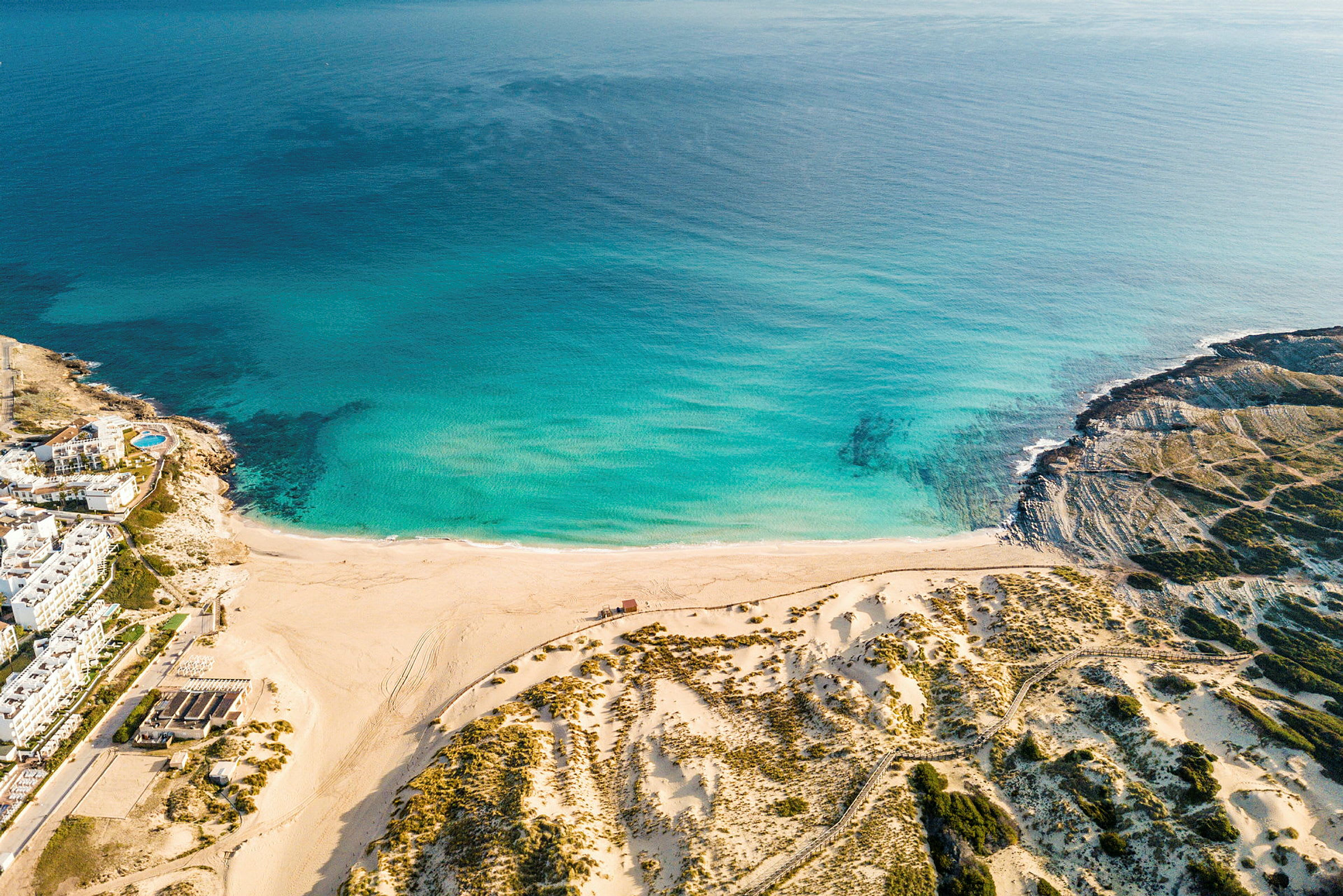 Flygbild över en strand med turkost vatten och sanddyner, omgiven av kustgröna områden. (AI-genererad text)
