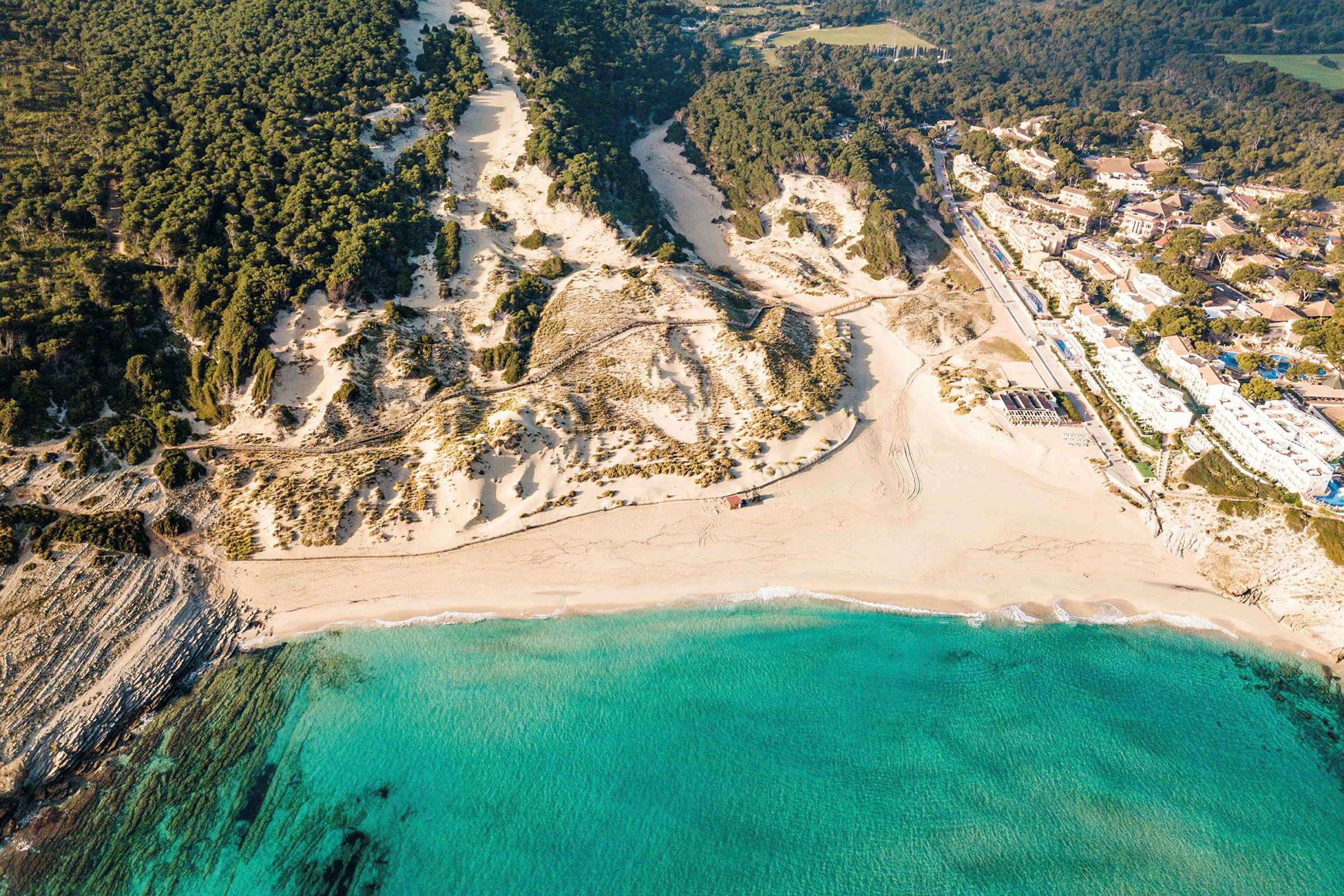Flygbild över en sandstrand med turkost vatten, omgiven av klippor och tallskog. (AI-genererad text)