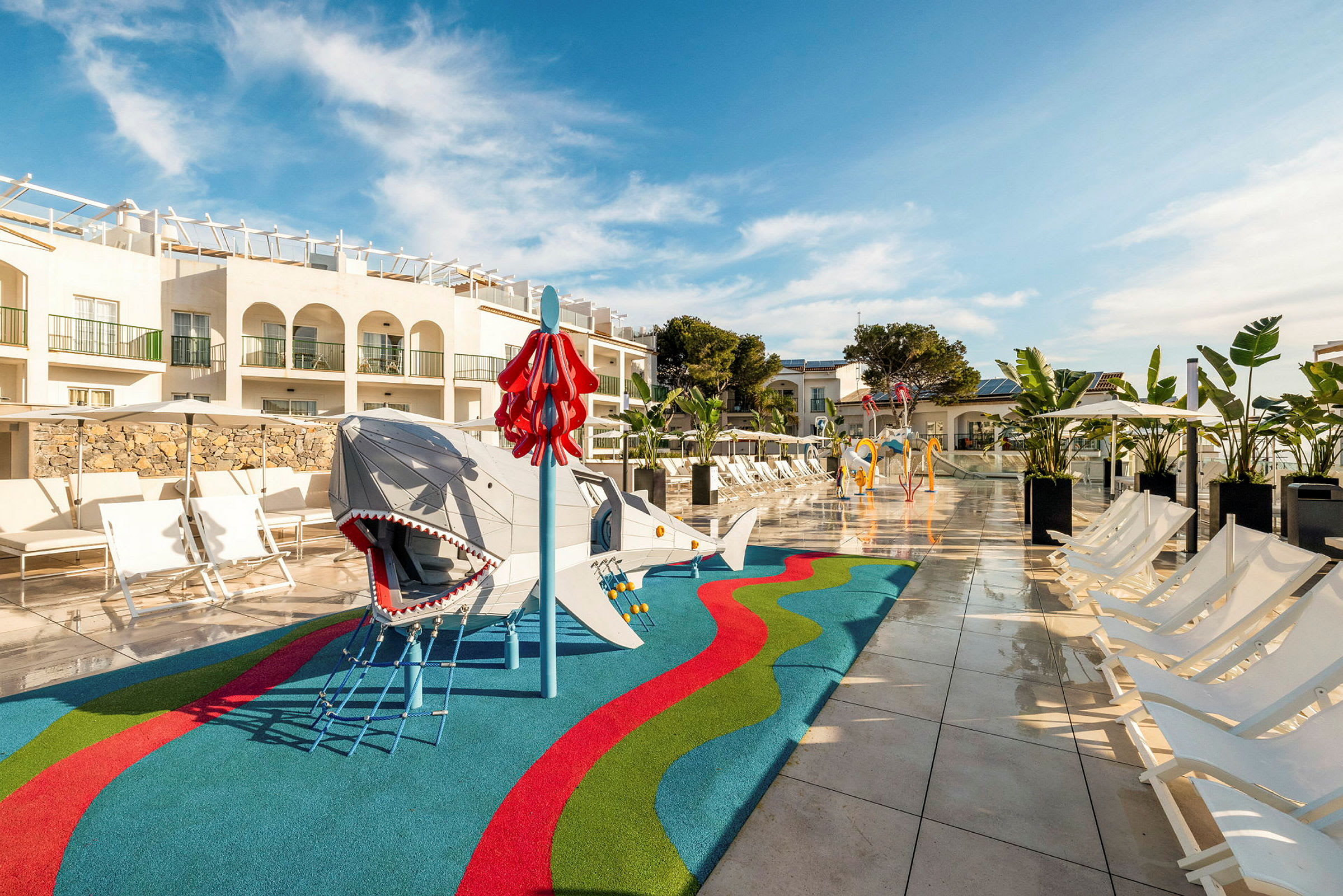 En lekplats med ett hajtema och färgglad markbeläggning ligger nära solstolar vid en pool, omgiven av semesterbyggnader med balkonger. (AI-genererad text)