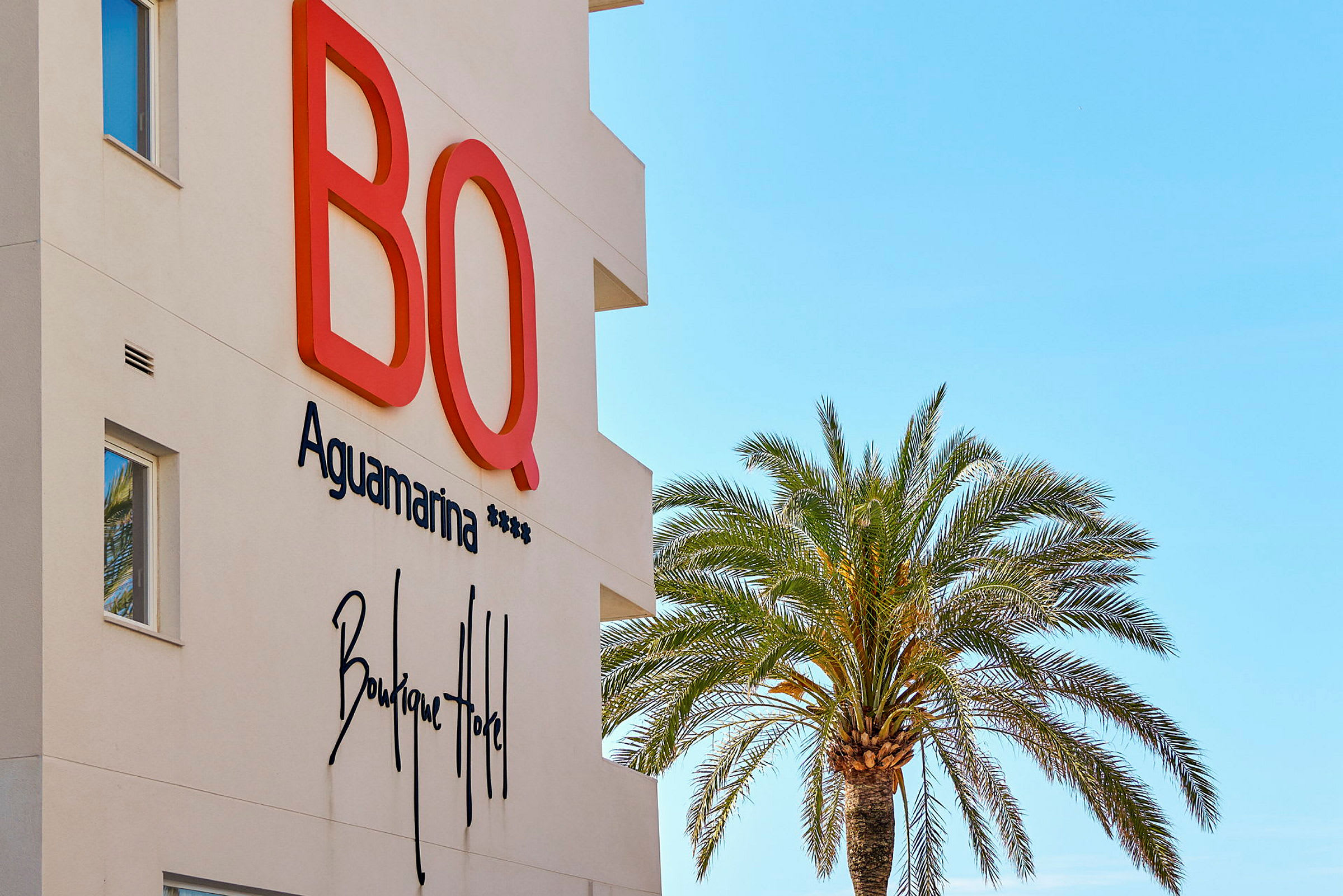 Fasad av BQ Aguamarina Boutique Hotel med en palm mot klar blå himmel. (AI-genererad text)