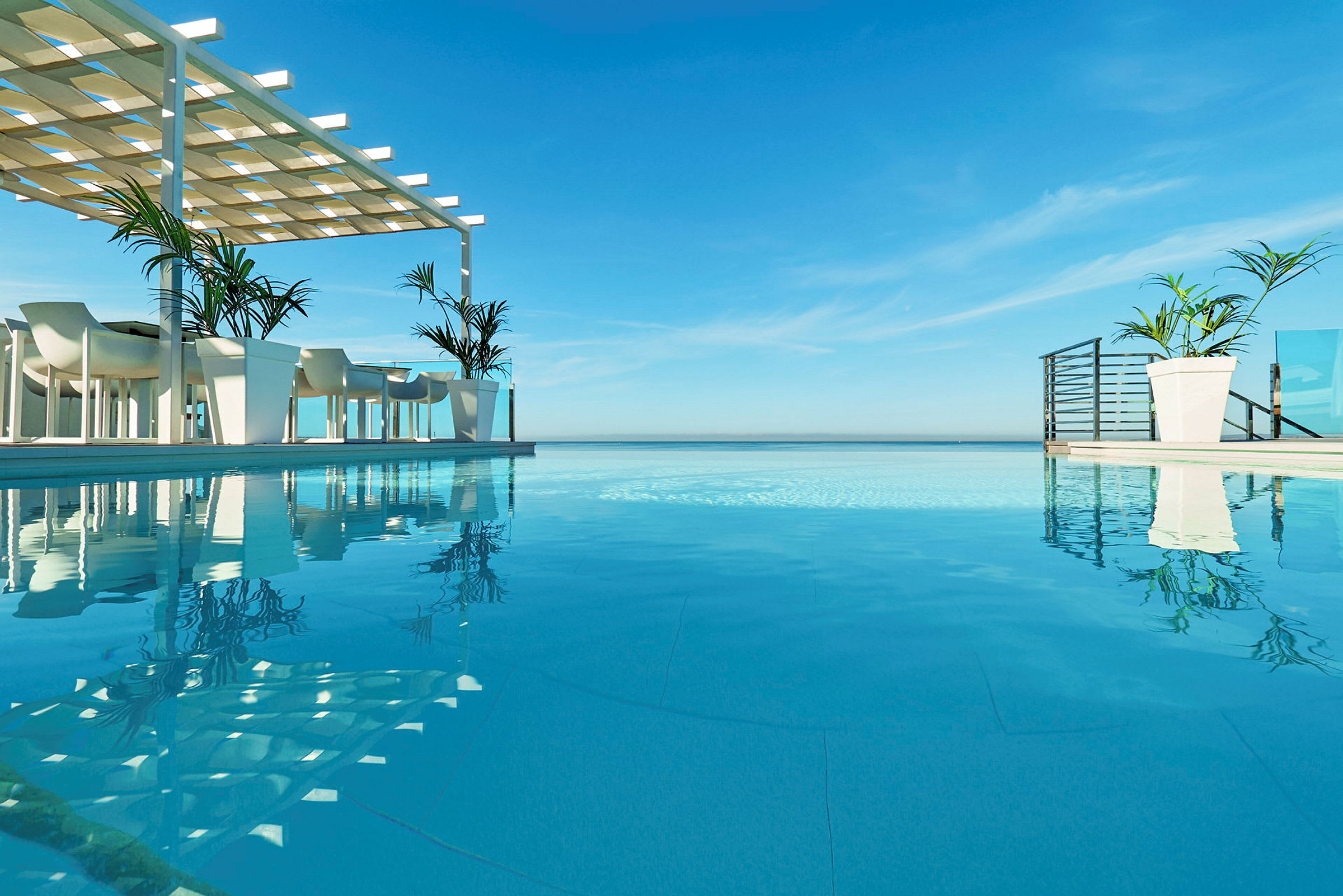 En infinitypool med utsikt över havet, med bord och stolar under en skuggstruktur bredvid poolen. (AI-genererad text)