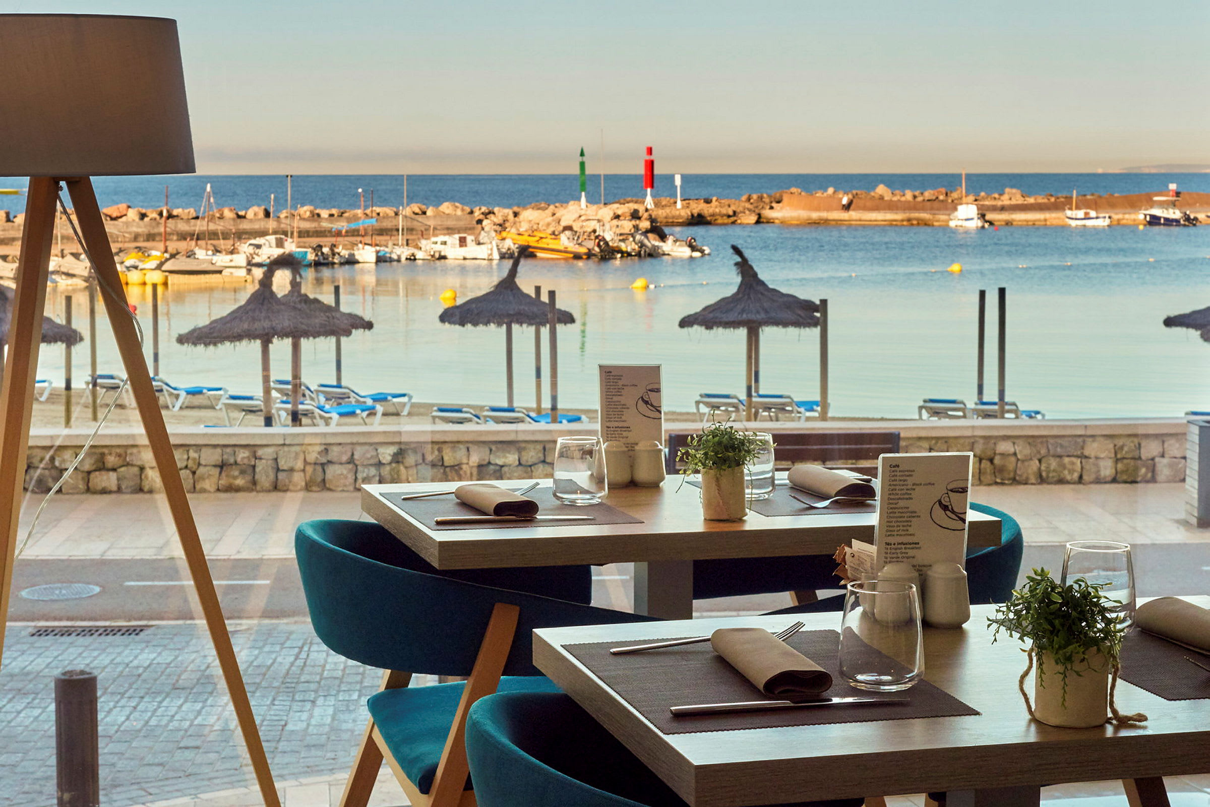 Utsikt från en restaurang mot en strand med parasoller och solstolar, samt en småbåtshamn med båtar. (AI-genererad text)
