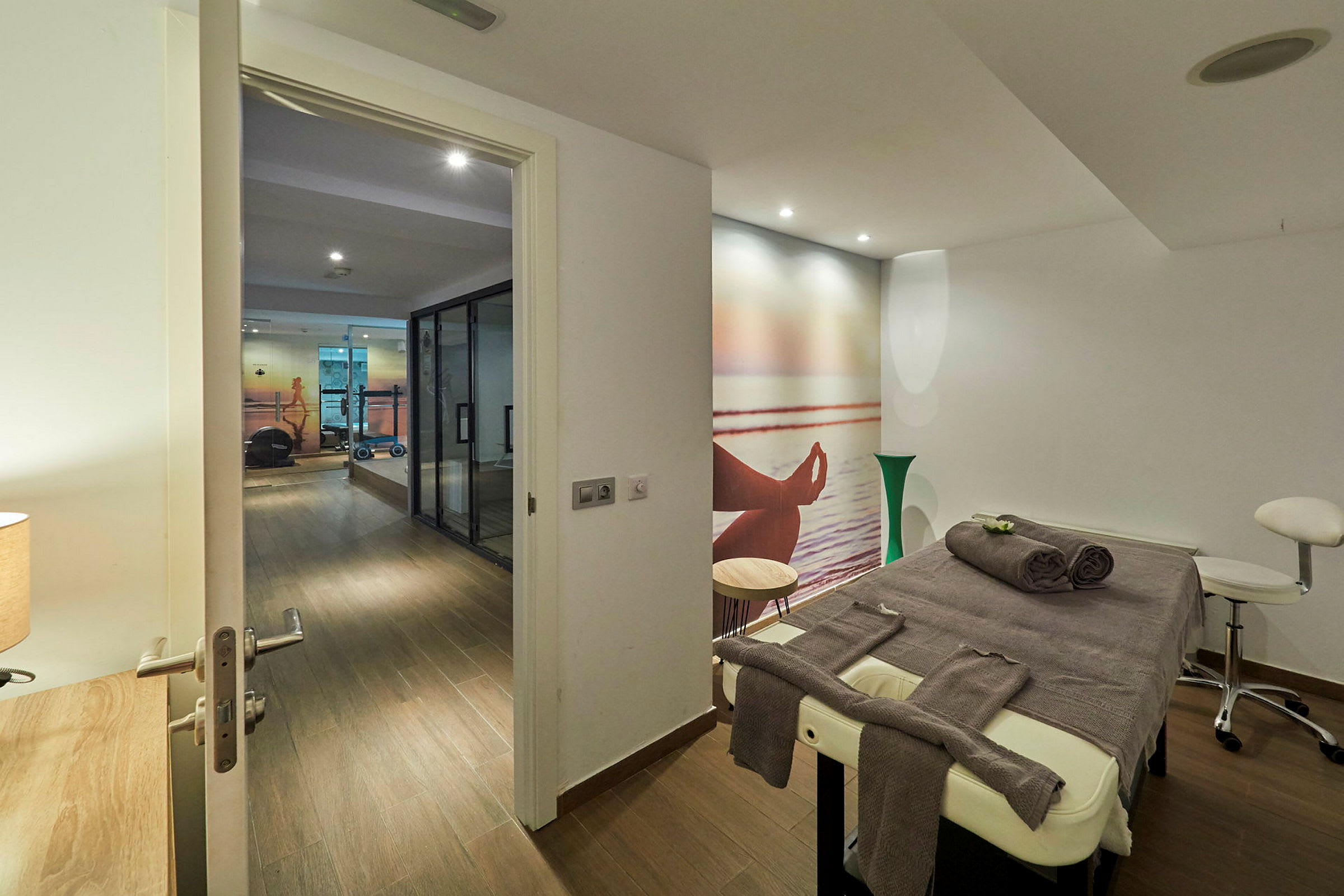 En massagebänk med handdukar i ett behandlingsrum med en väggmålning av ett mediterande motiv, bredvid ett gym. (AI-genererad text)