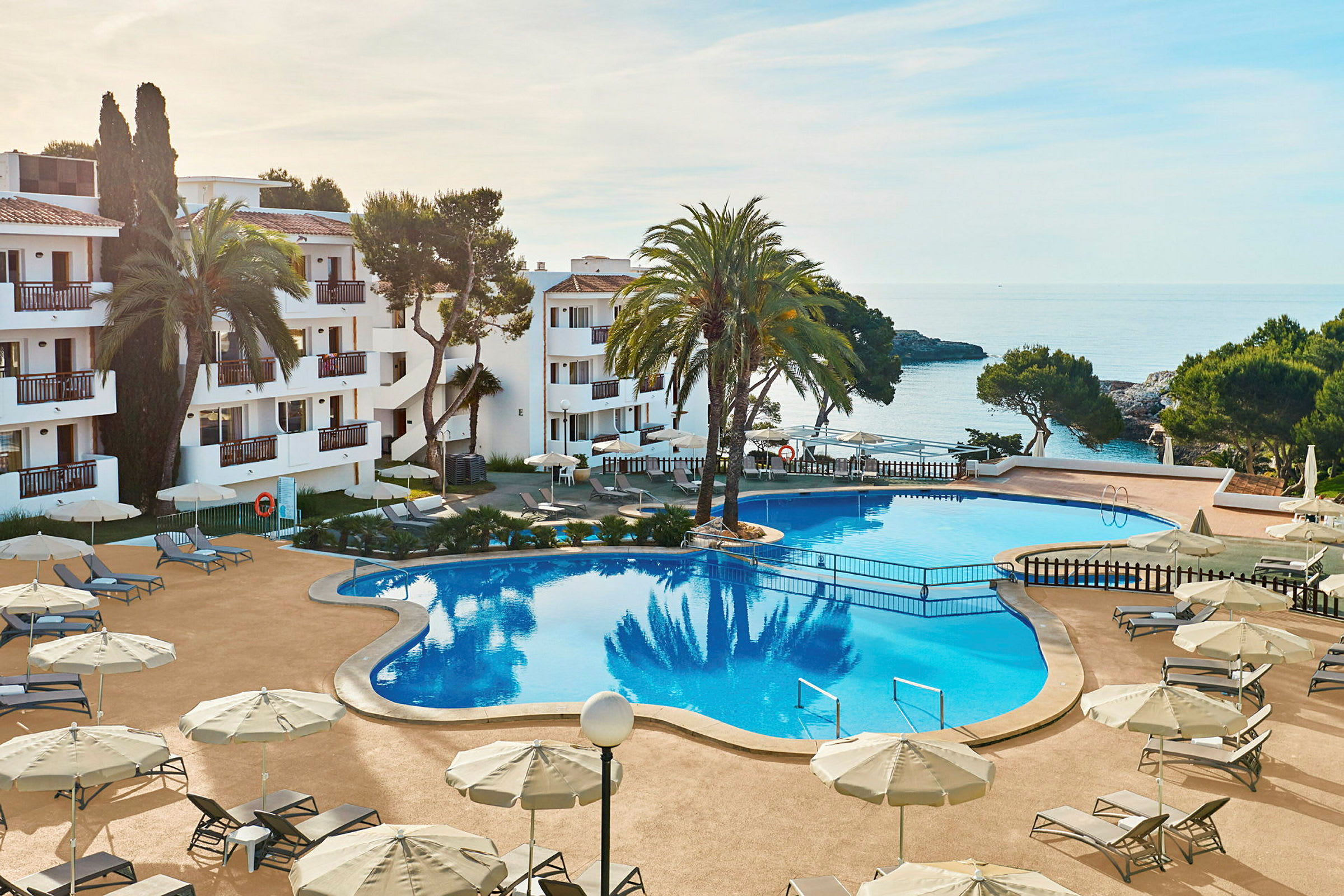 Bild av ett hotellområde med en stor pool, omgiven av solstolar och parasoller. I bakgrunden syns havet och delar av byggnaden. (AI-genererad text)