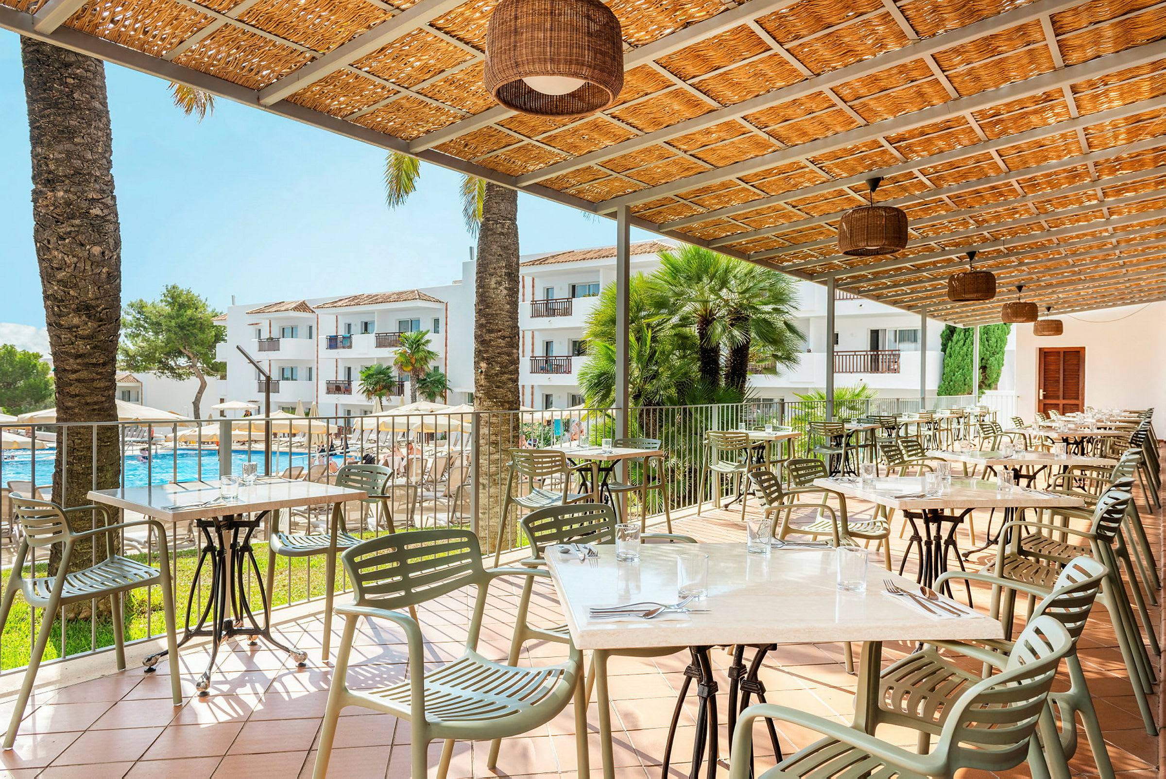 En utomhusrestaurang med bord och stolar under ett tak, med utsikt över en pool och hotellbyggnader i bakgrunden. (AI-genererad text)