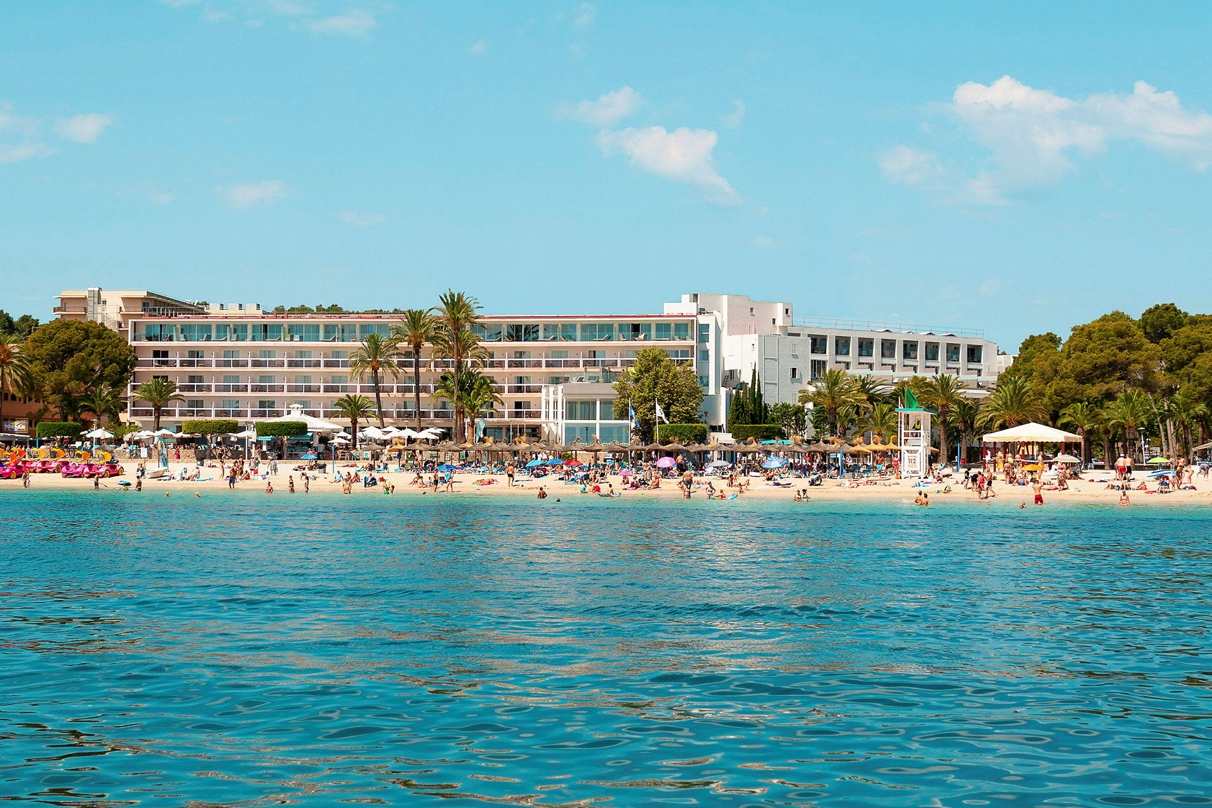 Ett hotell vid en strand med palmer och badande människor. (AI-genererad text)