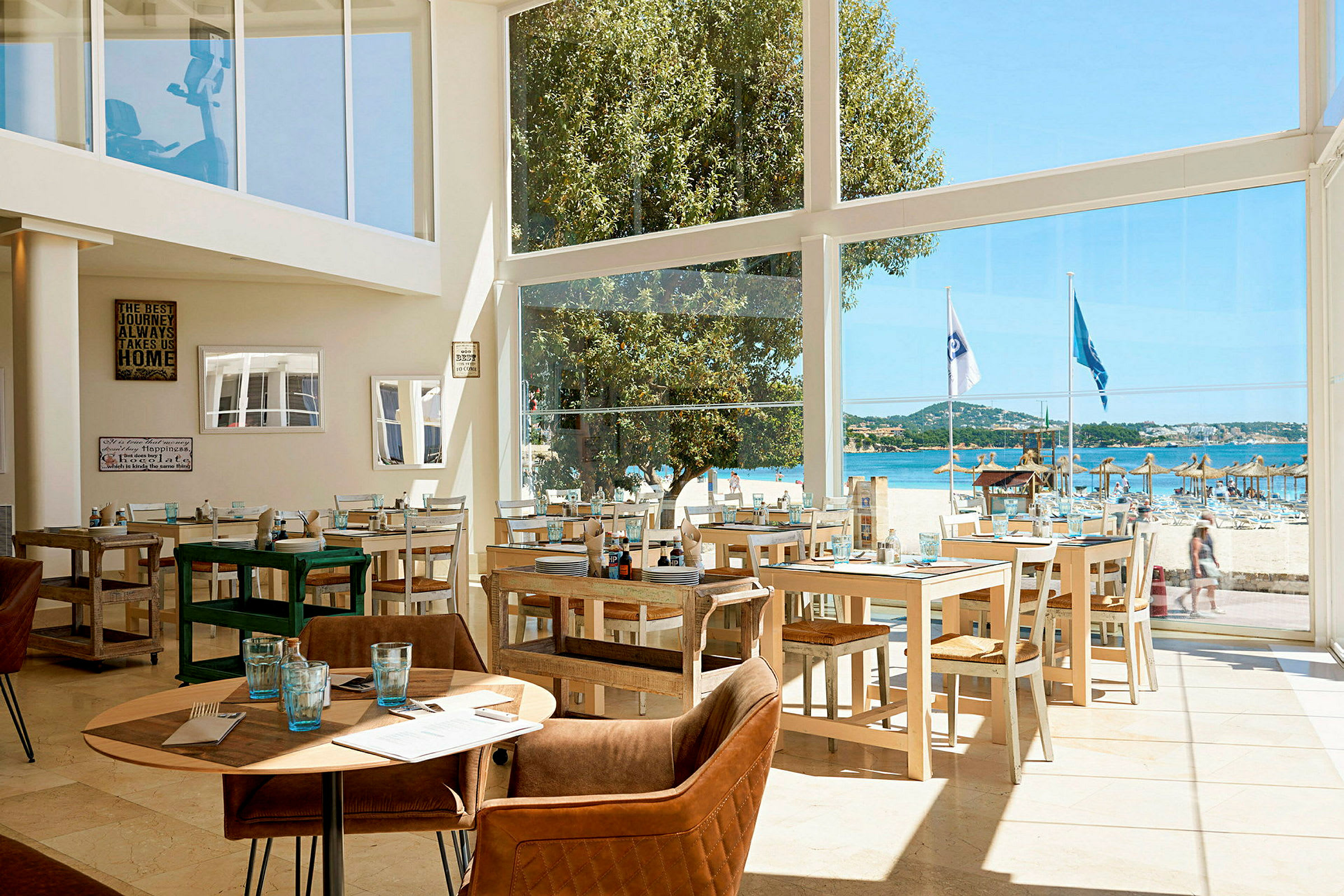 En restaurang med glasväggar som ger utsikt över en strand med parasoll och blått hav. (AI-genererad text)