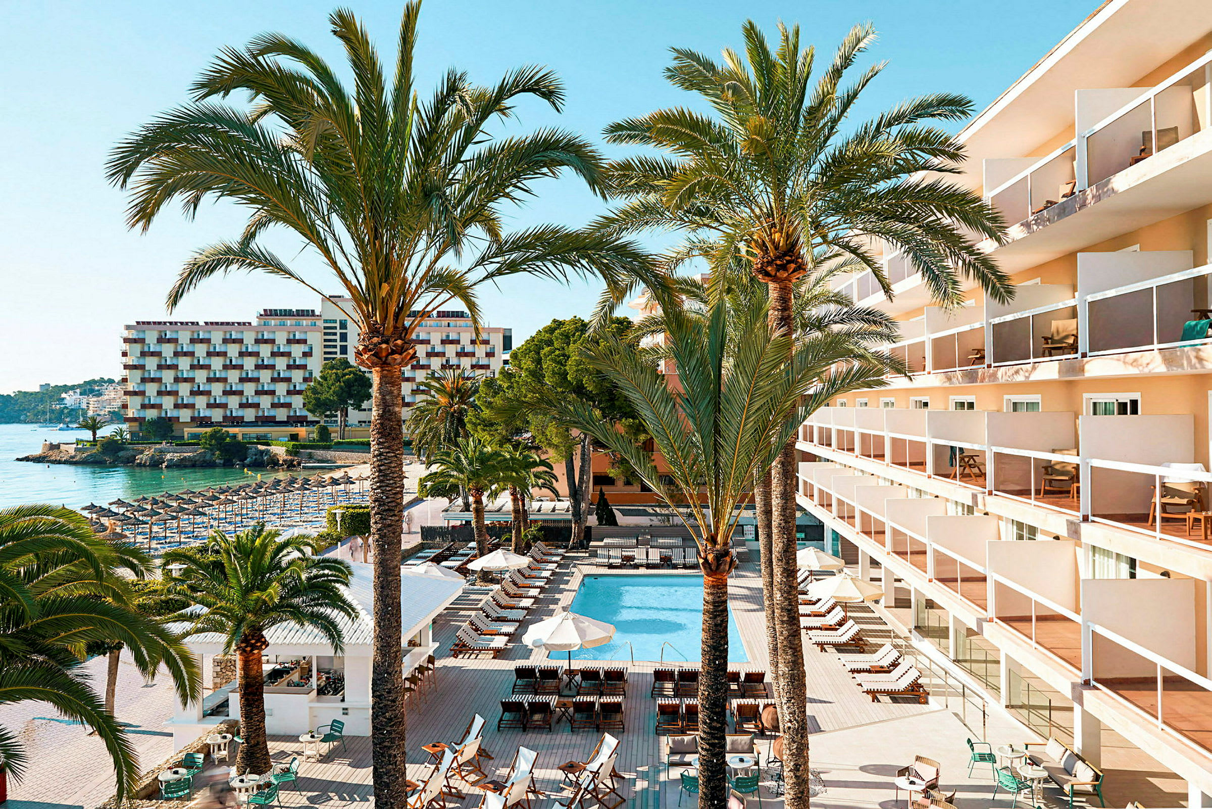 Bild av ett hotellområde med en pool, solstolar, parasoller och omgivande byggnader. Flera palmer syns i förgrunden. (AI-genererad text)