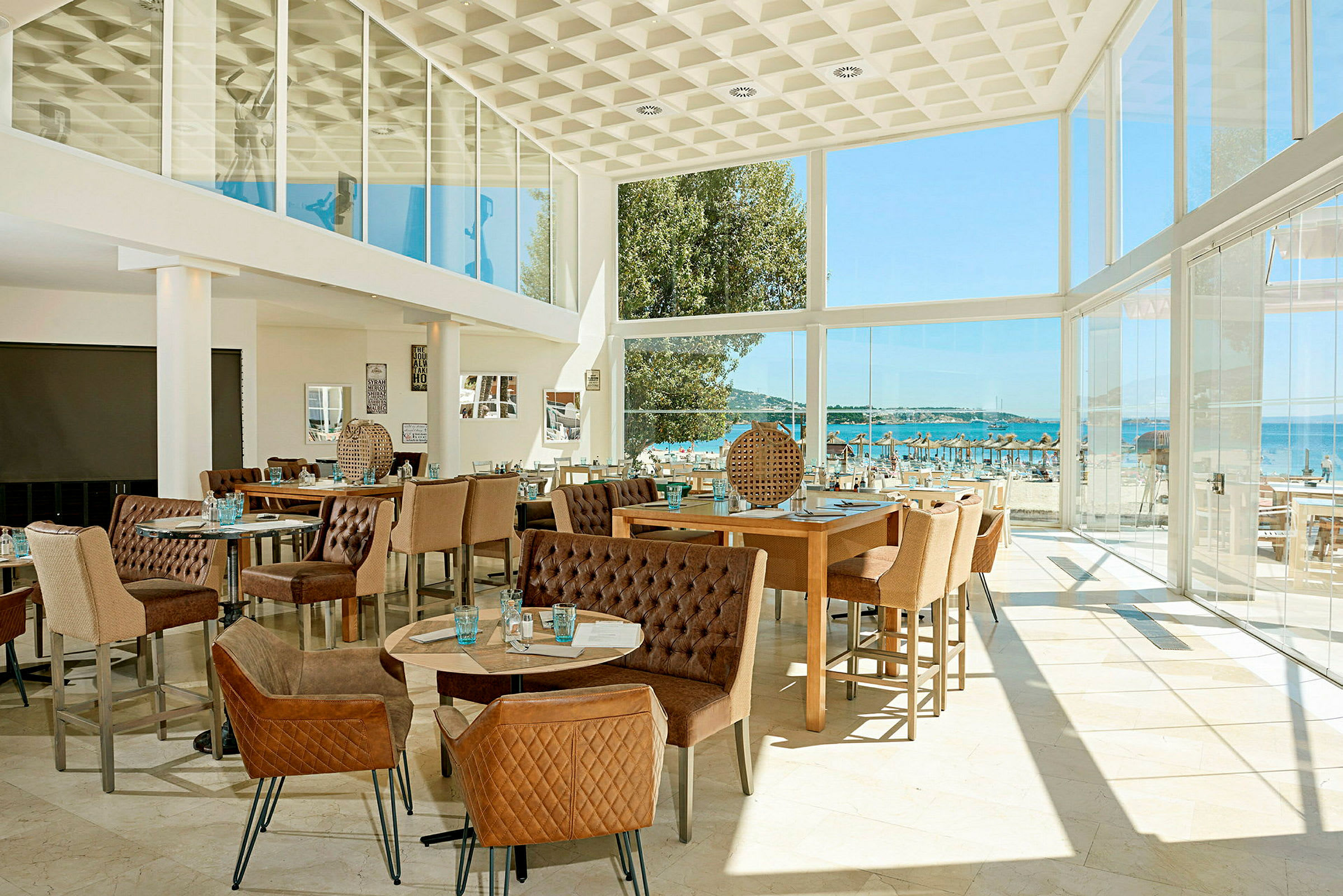 Ett modernt inrett restaurangutrymme med stora glasfönster som erbjuder utsikt mot en strand och havet utanför. (AI-genererad text)