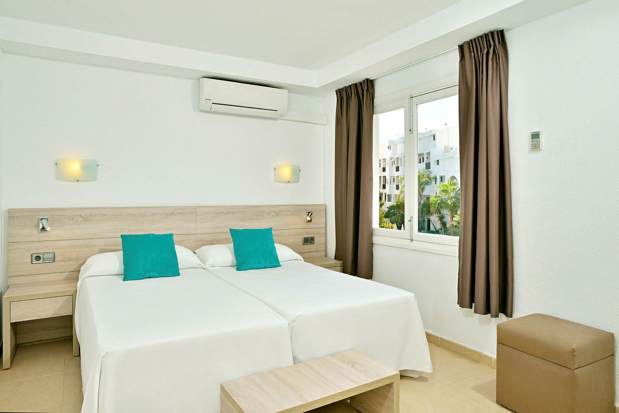 Ett hotellrum med två enkelsängar intill varandra, vit inredning och turkosa kuddar, samt ett fönster med utsikt. (AI-genererad text)