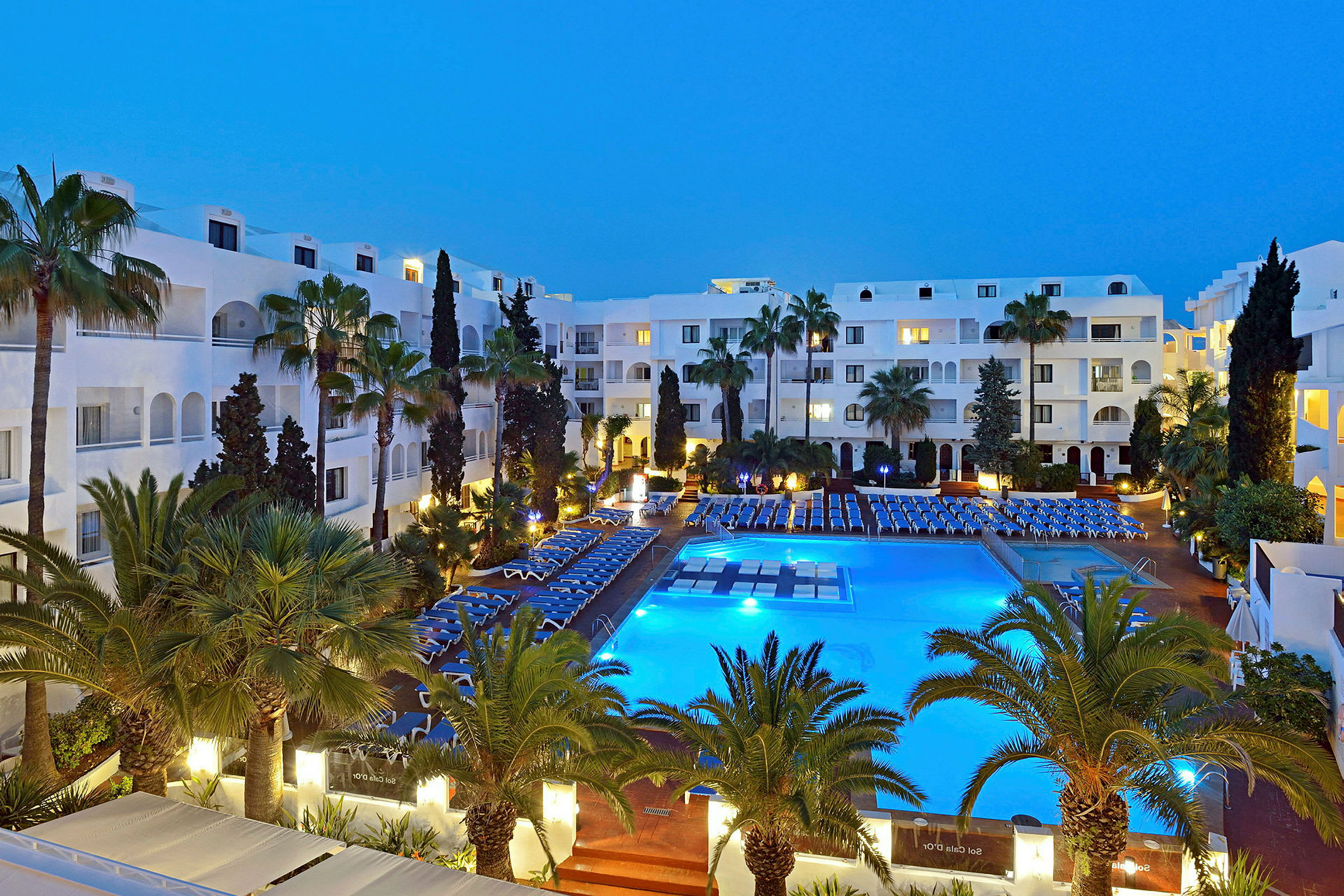 Ett hotellområde på kvällen med en upplyst pool, omgivande stolar och gröna palmer. (AI-genererad text)