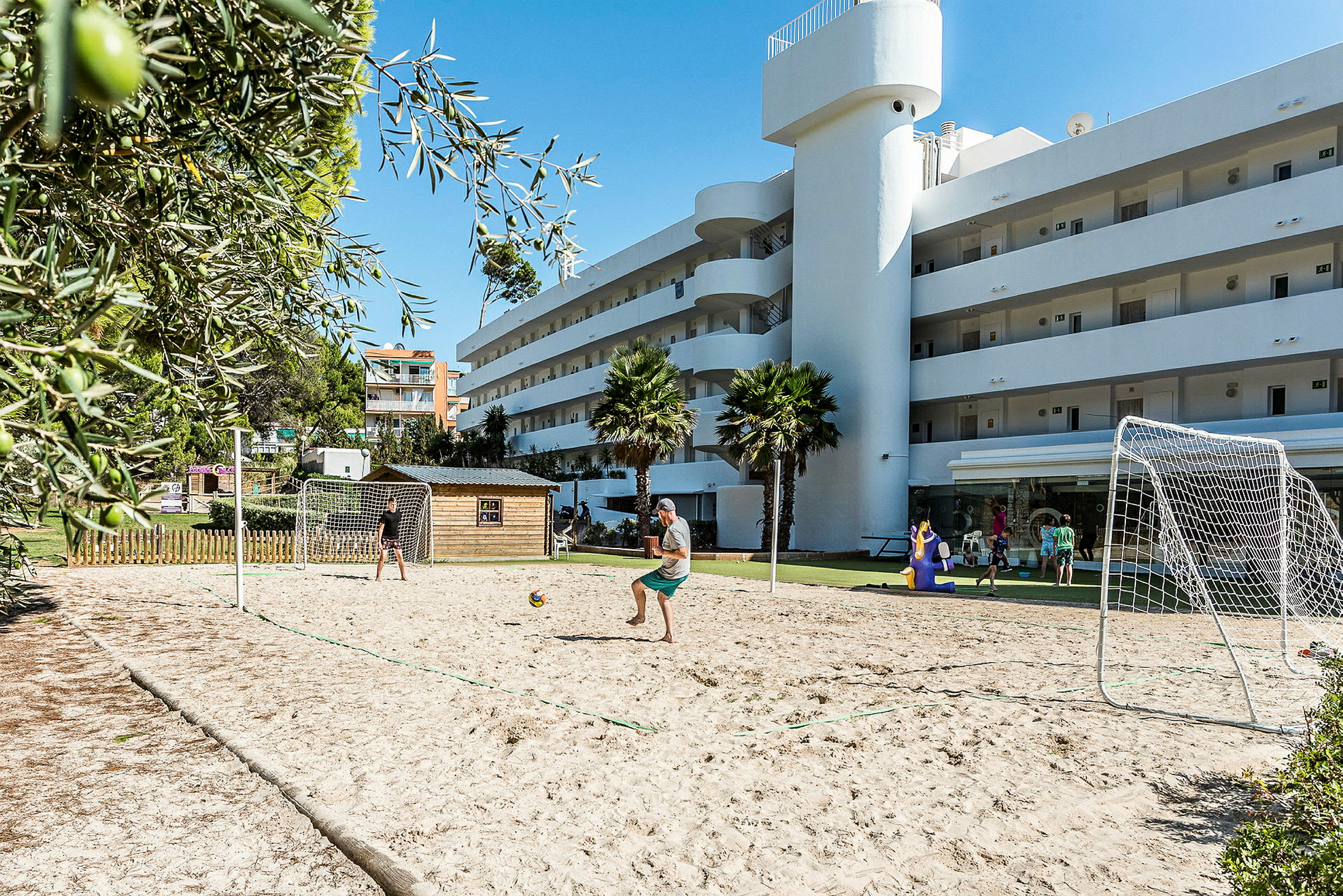 En utomhus plan med sand för spel såsom fotboll framför en vit byggnad. (AI-genererad text)