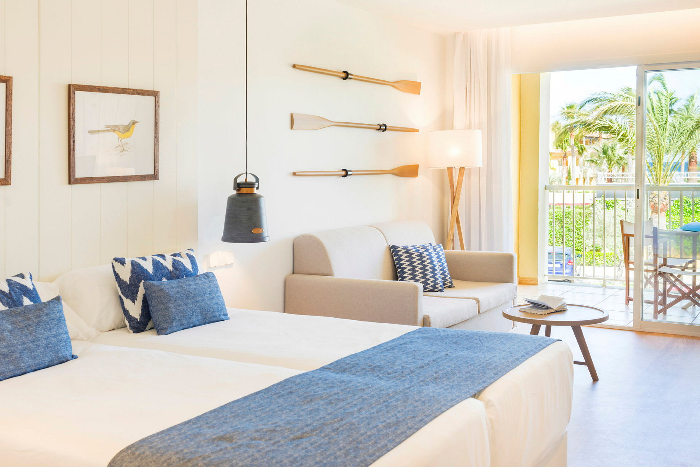 Ett hotellrum med två sängar, en soffa, en lampa, dekorerade paddlar på väggen och en balkong med utsikt. (AI-genererad text)