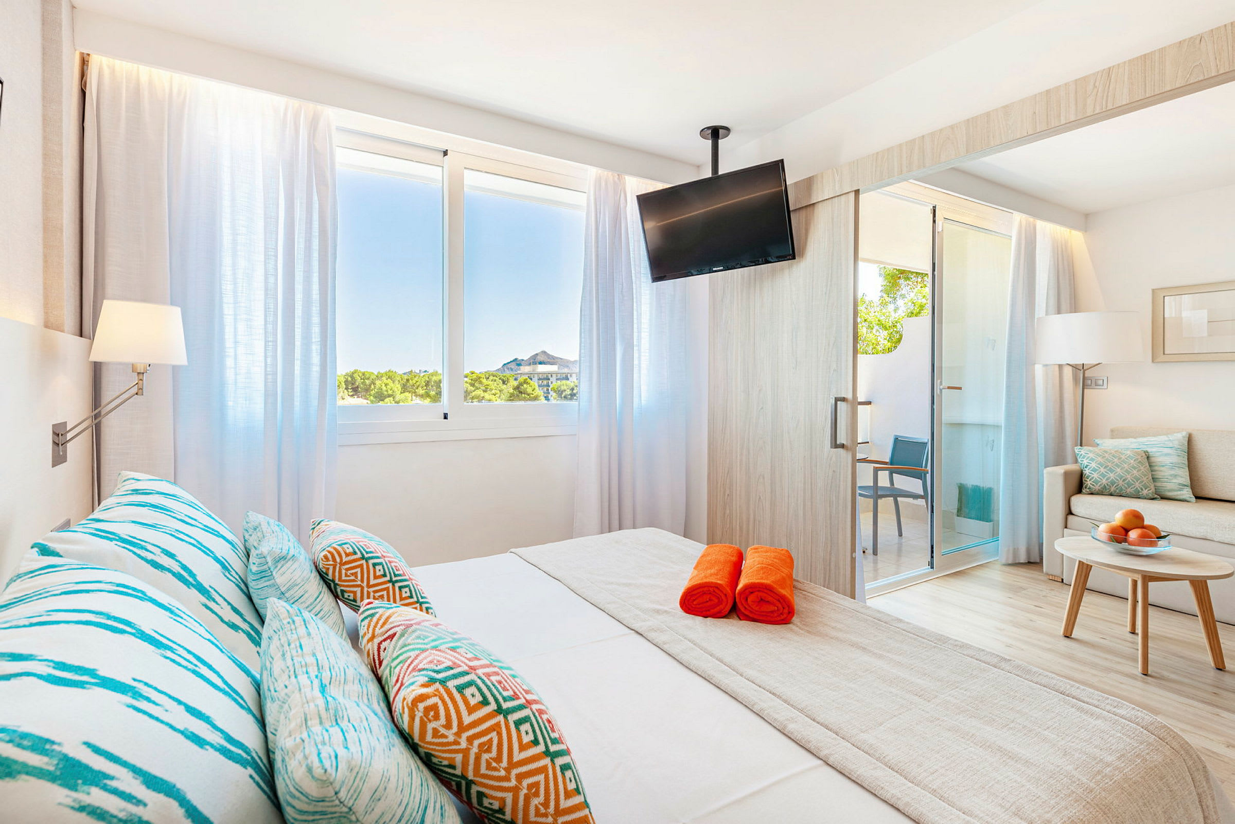 Ljust hotellrum med säng, färgglada kuddar, och orange handdukar. (AI-genererad text)