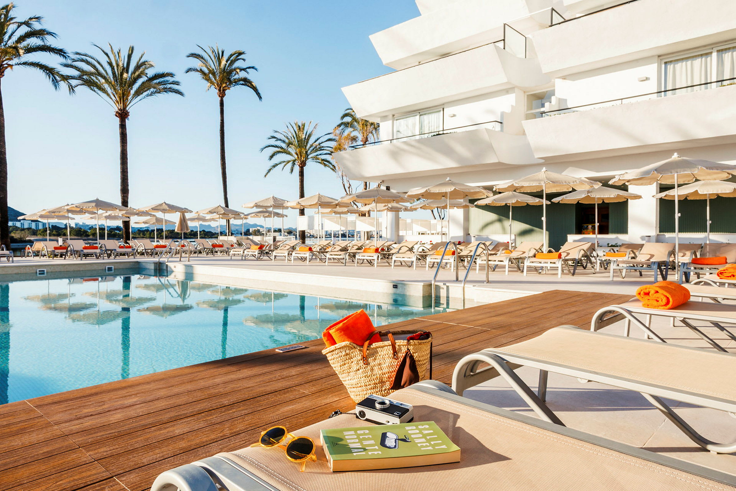 Bild av en pool med solstolar, solparasoller och omgivande palmträd. Ett vitt hotell syns i bakgrunden. (AI-genererad text)