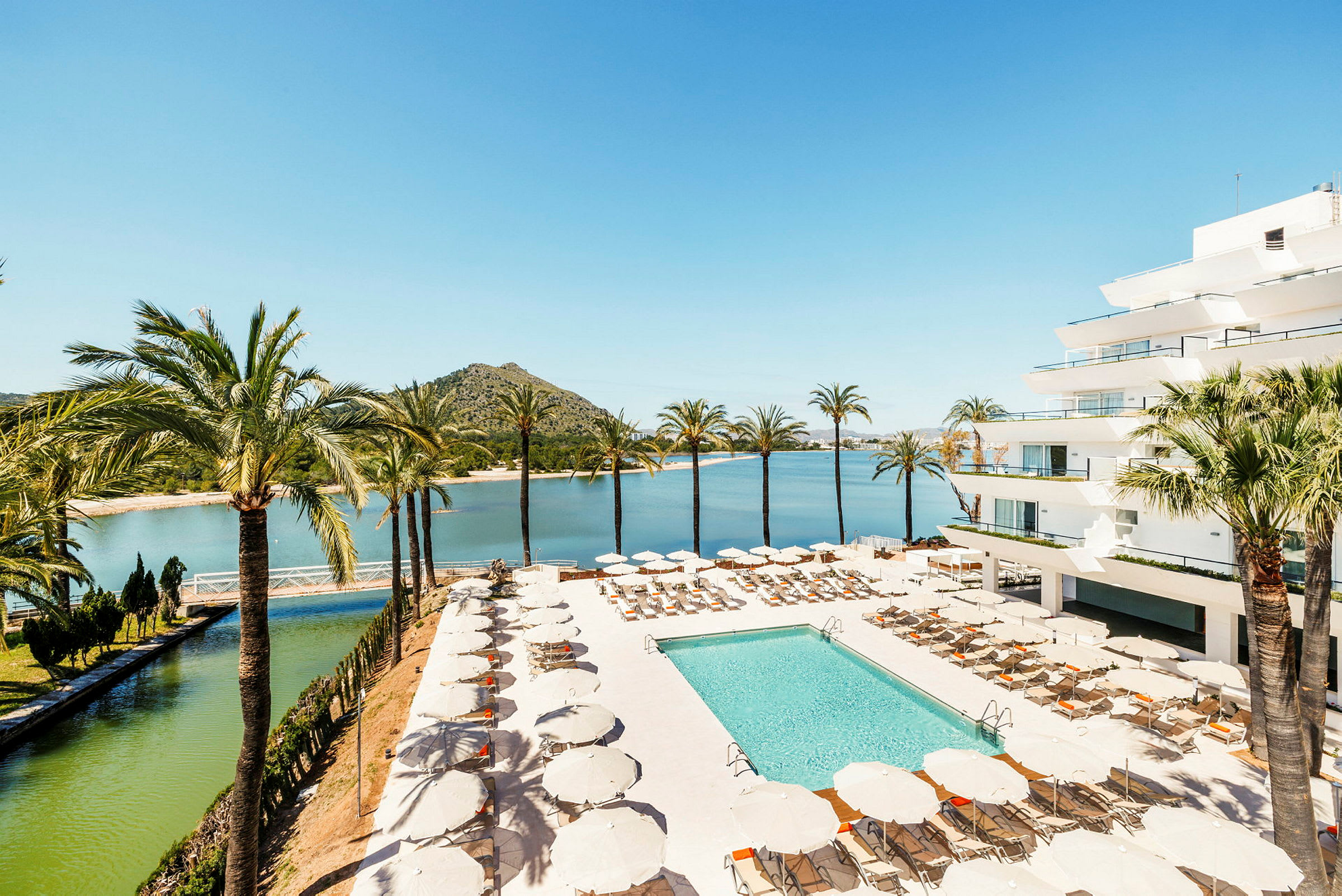 Bild på en hotellpool med solstolar och parasoller omgivna av palmer, med en vy över en sjö och grönklädda kullar i bakgrunden. (AI-genererad text)