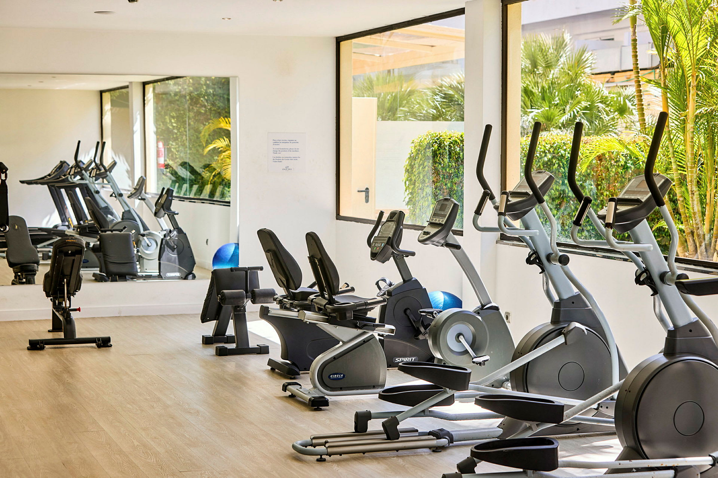 Ett ljust gym med träningsmaskiner inklusive motionscyklar och crosstrainers på ett trägolv, omringat av fönster med utsikt över grönska och spegel på ena väggen. (AI-genererad text)