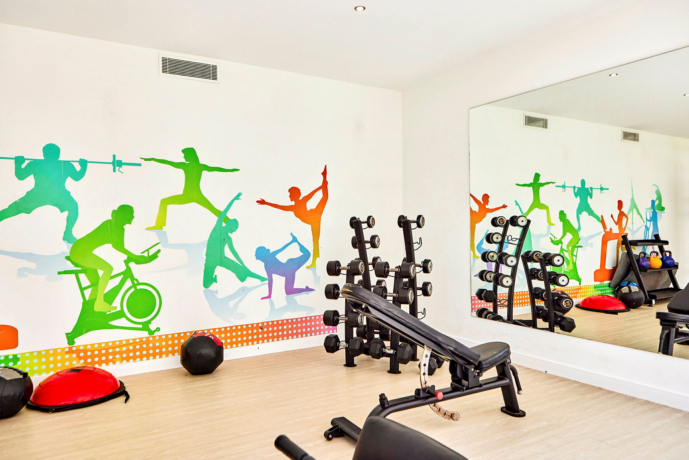Ett gym med träningsutrustning och en färgglad väggmålning föreställande silhuetter av människor som tränar. (AI-genererad text)