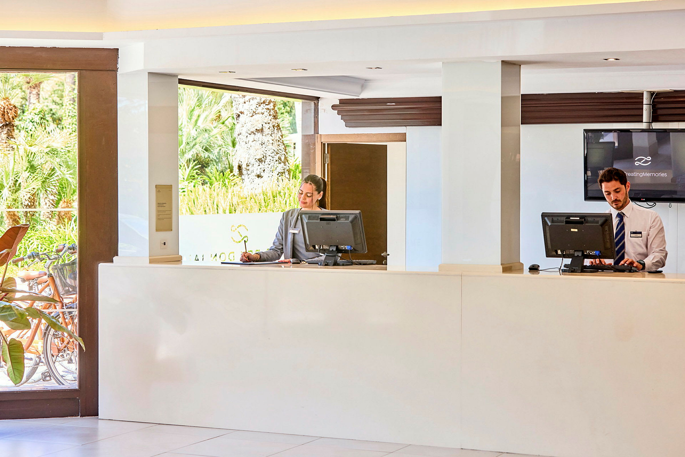 Två receptionister bakom en receptionsdisk i ett hotell med grönska utanför. (AI-genererad text)
