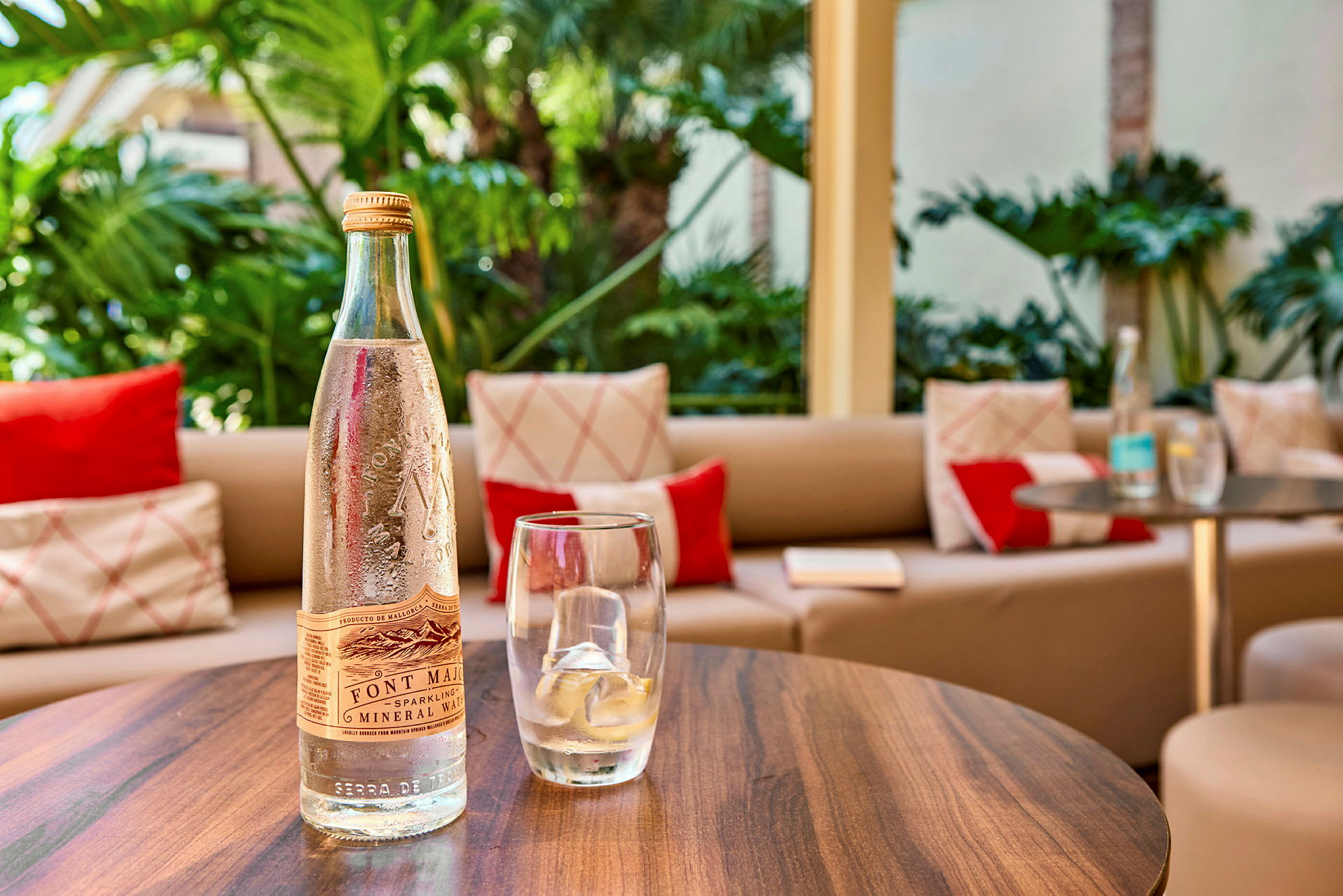 Ett bord med en flaska mineralvatten, ett glas med is, och i bakgrunden syns en sittplats med kuddar samt gröna växter. (AI-genererad text)