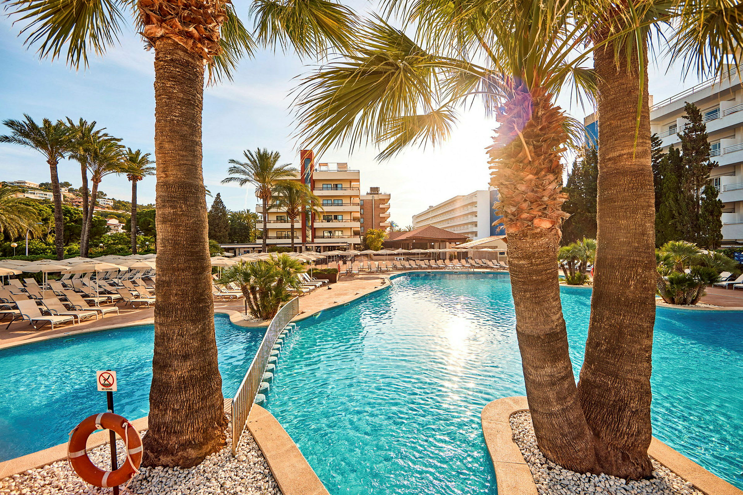 Hotellpool med omgivande solstolar och palmer i förgrunden. (AI-genererad text)