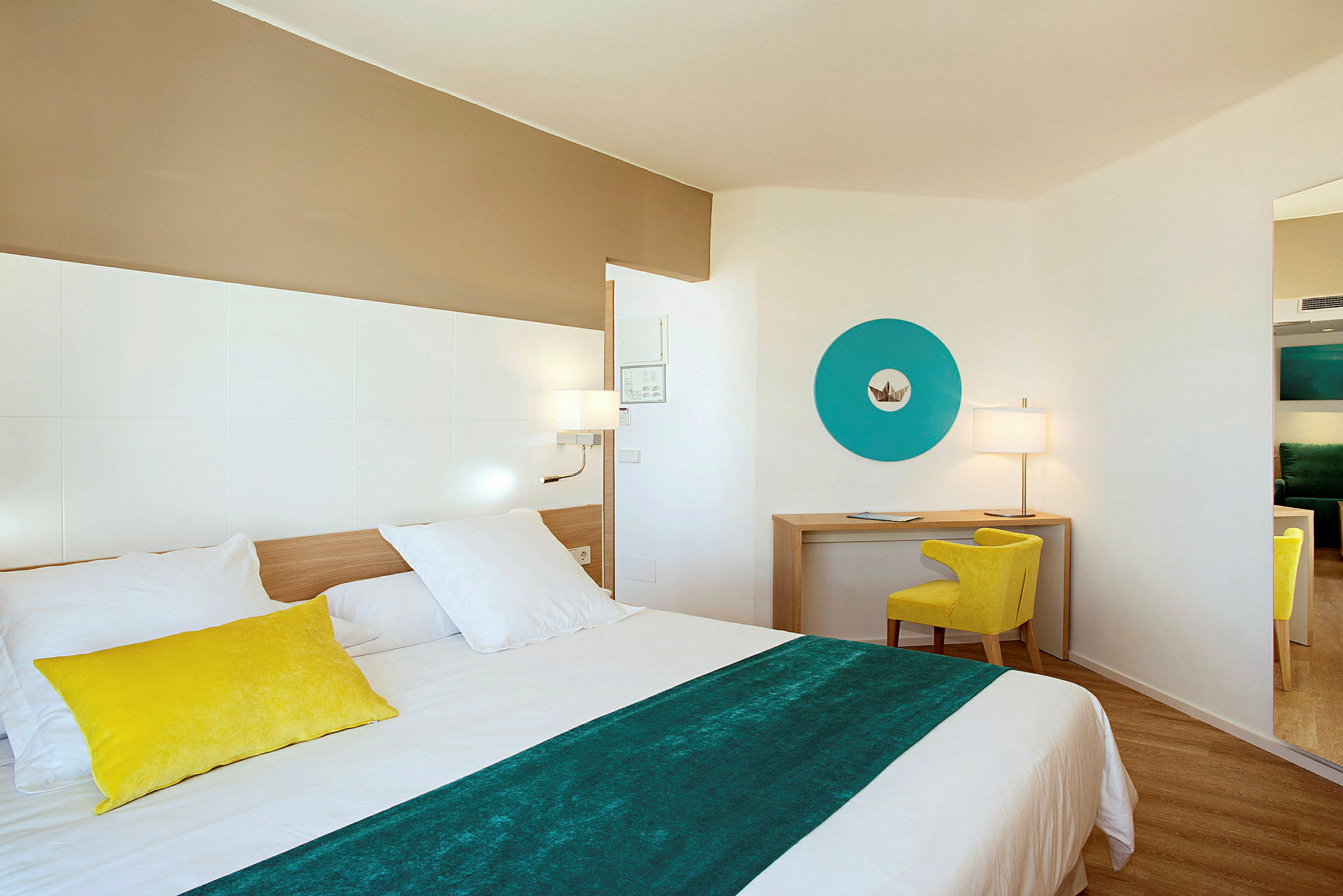 Ett modernt hotellrum med en säng i förgrunden, en skrivbordshörna med en gul stol och en blå dekorativ väggdetalj. (AI-genererad text)