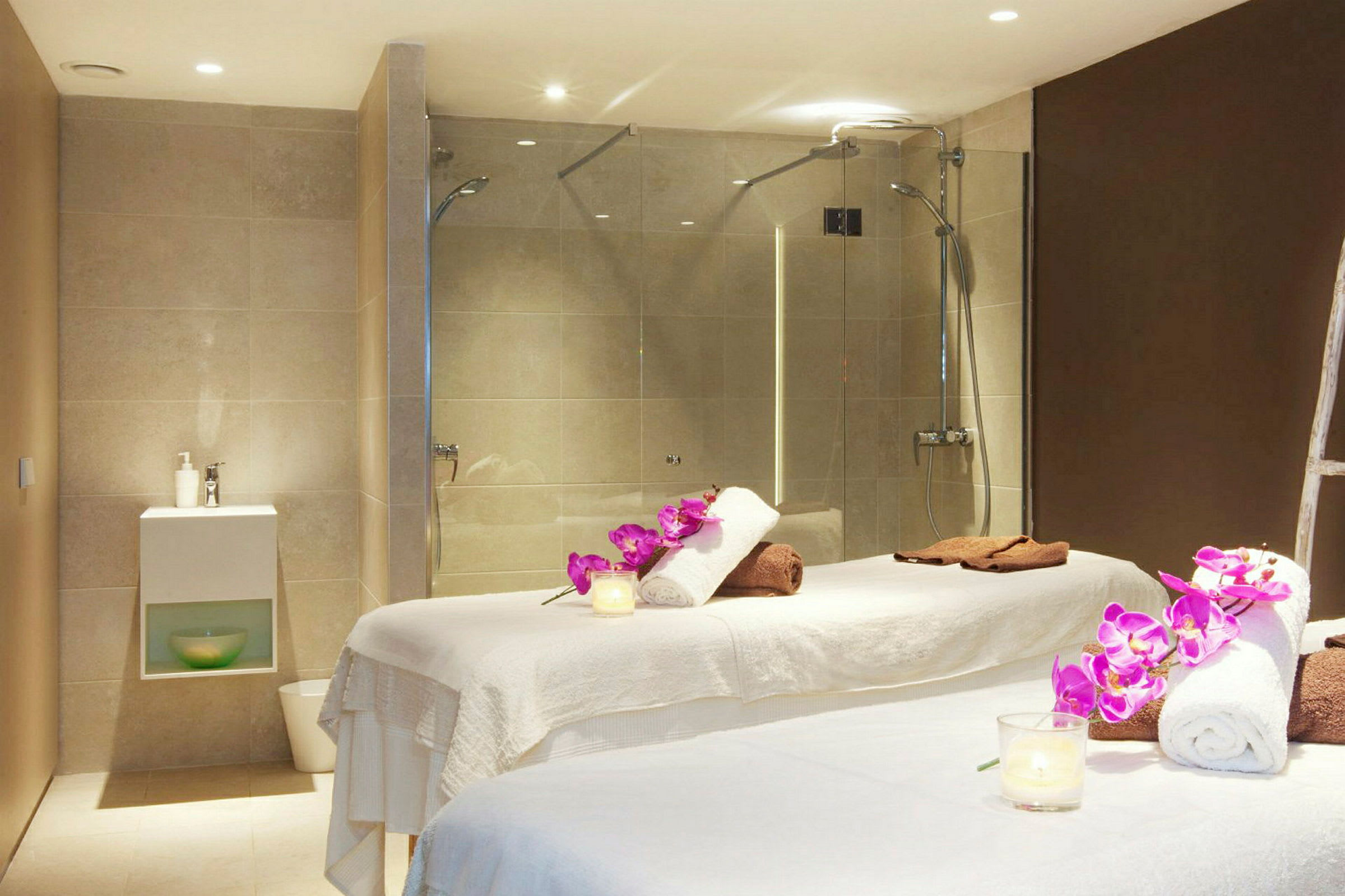 Bild av ett spa med två massagebänkar, handdukar, orkidéer och en dusch i bakgrunden. (AI-genererad text)