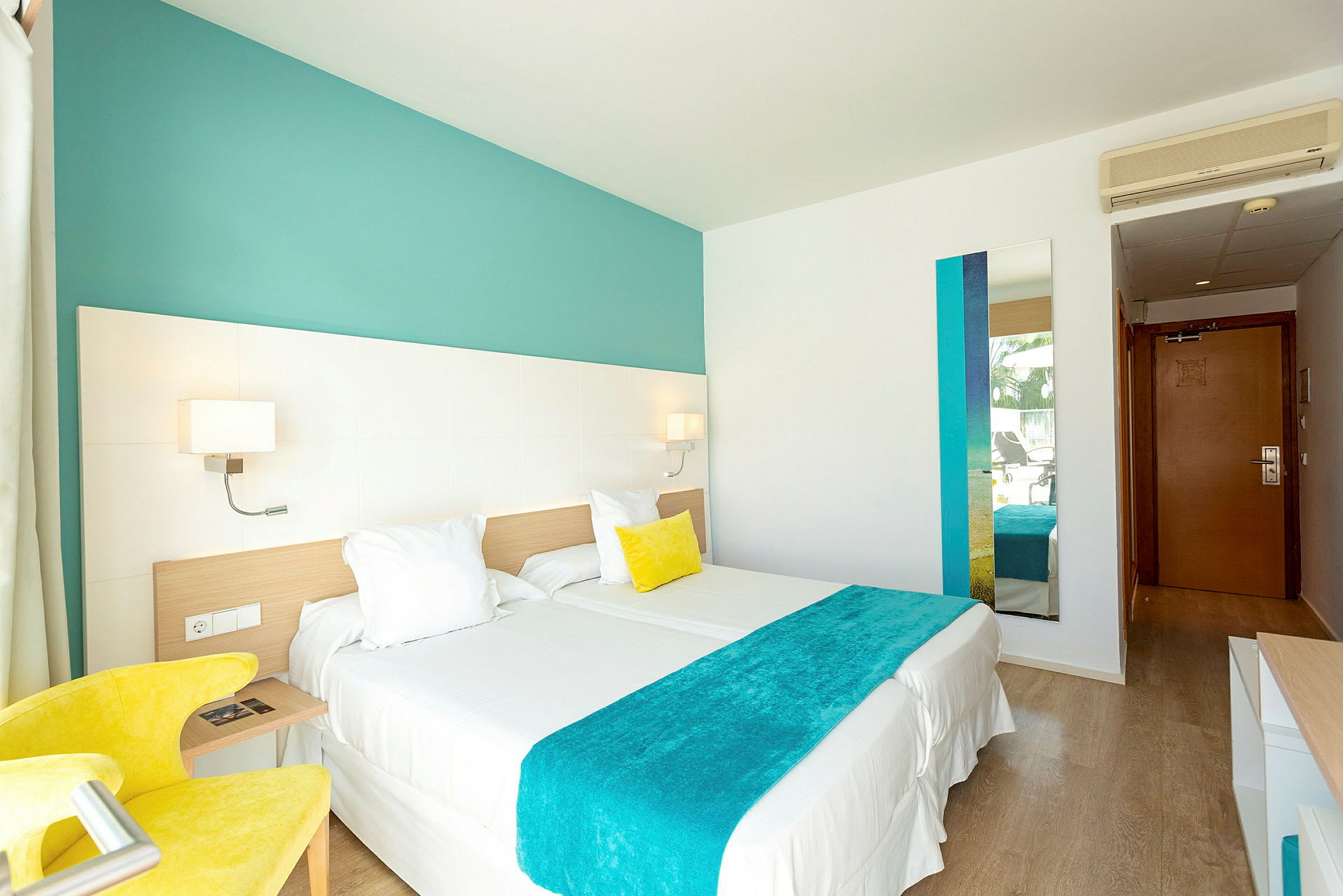 Ett ljust hotellrum med två enkelsängar som är sammanställda, en gul fåtölj, väggdekoration i turkosa nyanser och en spegel. En dörr till entrén ses i bakgrunden. (AI-genererad text)