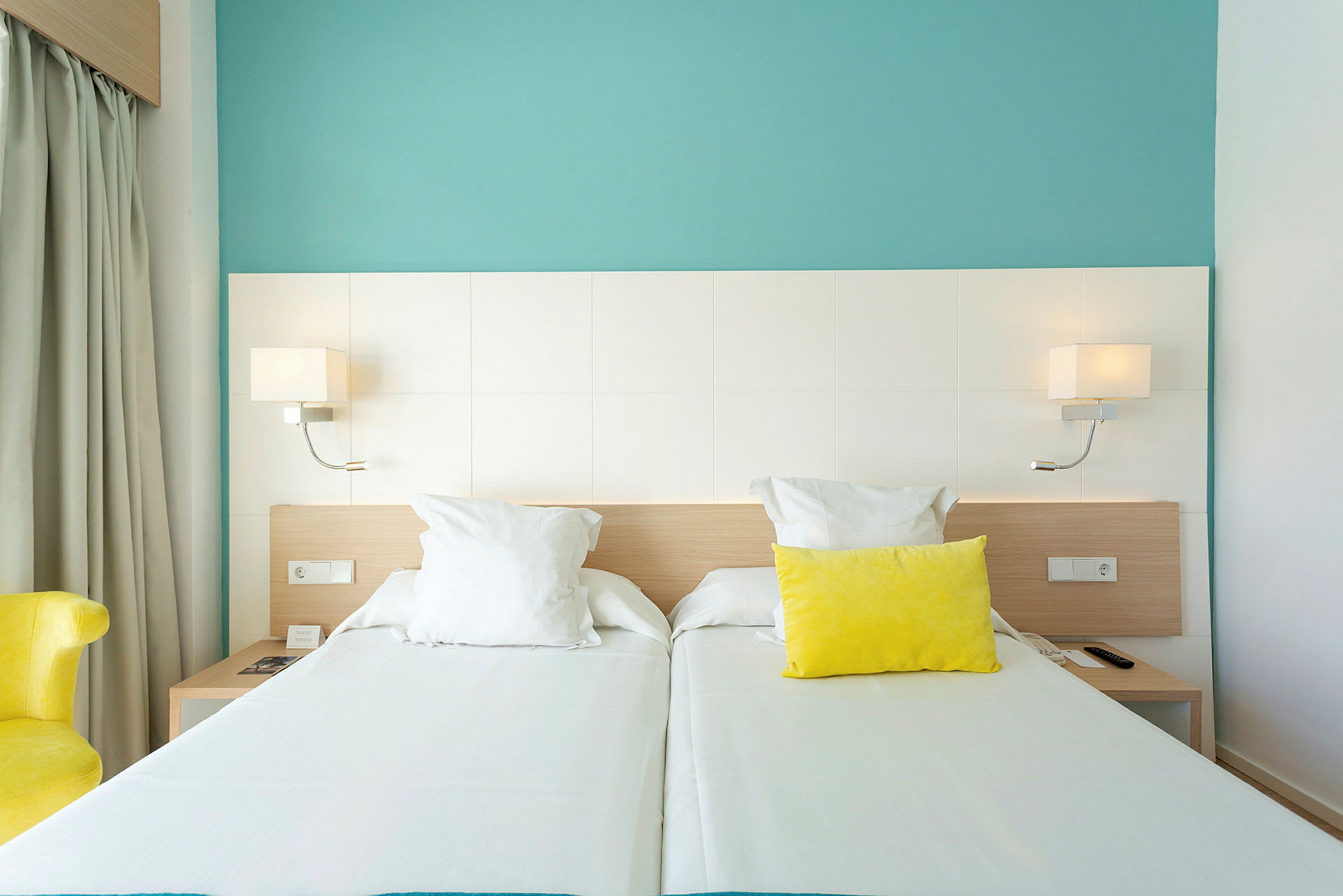 Ett hotellrum med två enkelsängar, en gul kudde och en ljusblå vägg. (AI-genererad text)