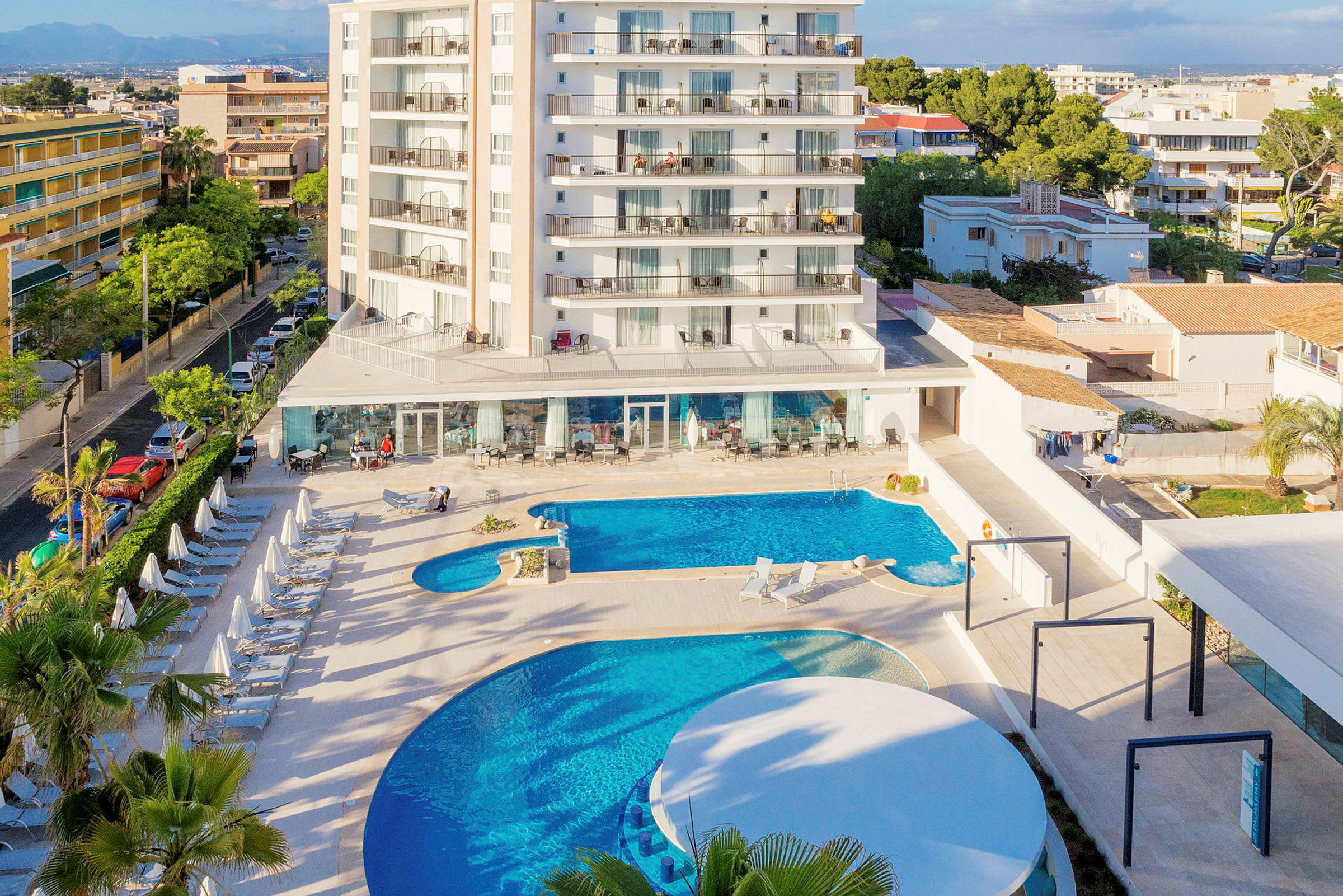 Hotell med poolområde och solstolar, omgivna av byggnader och träd. (AI-genererad text)
