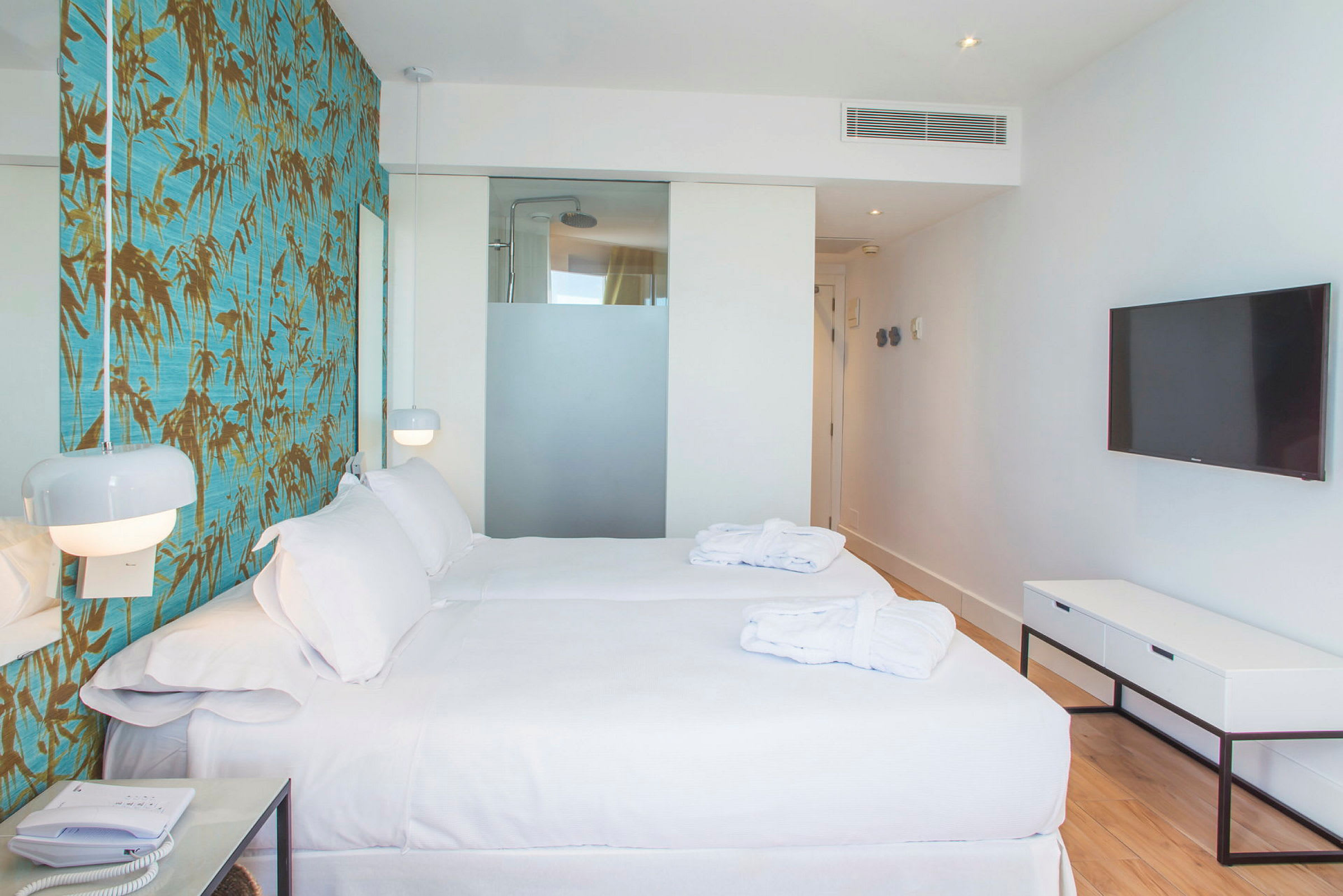 Ett hotellrum med två enkelsängar, vit sängkläder, en vägg med mönstrad tapet, en platt-TV och en glasvägg som delar av badrummet. (AI-genererad text)