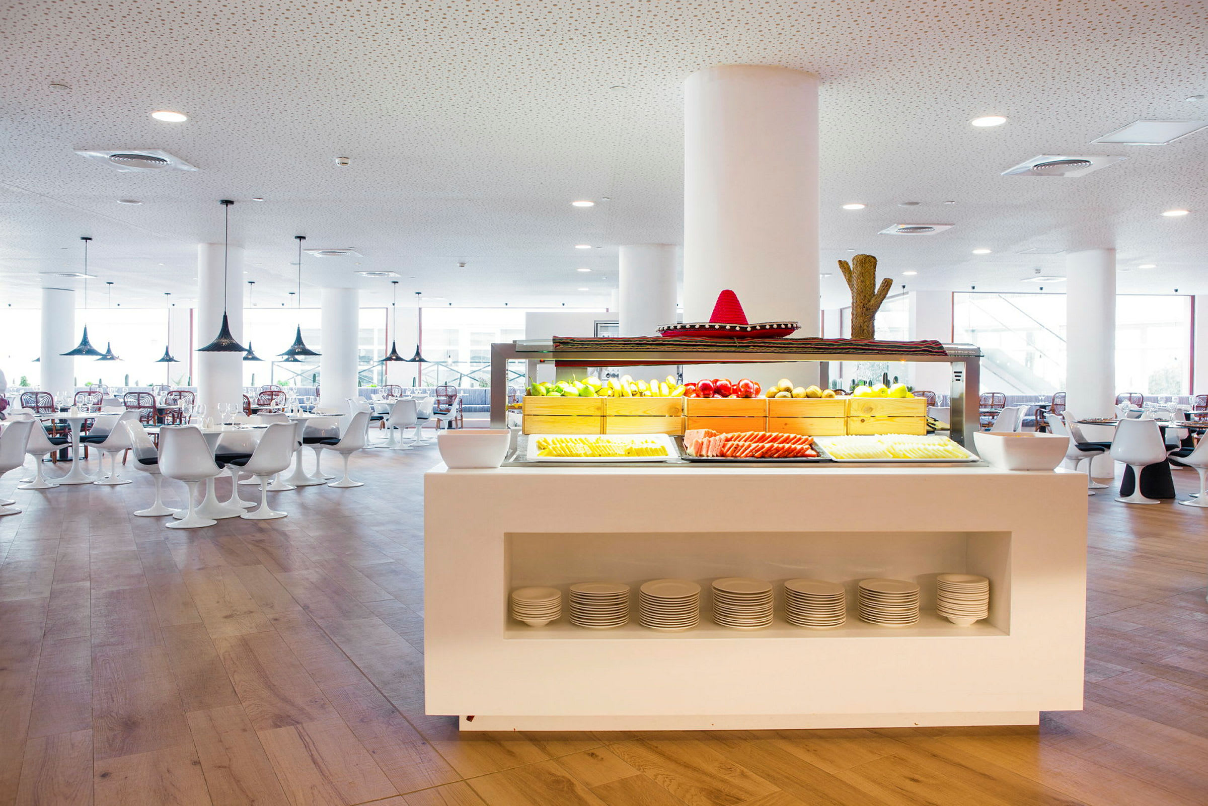 Ett restaurangområde med en vit bufféstation fylld med färsk frukt och grönsaker, omgiven av moderna vita bords- och sittmöbler. (AI-genererad text)