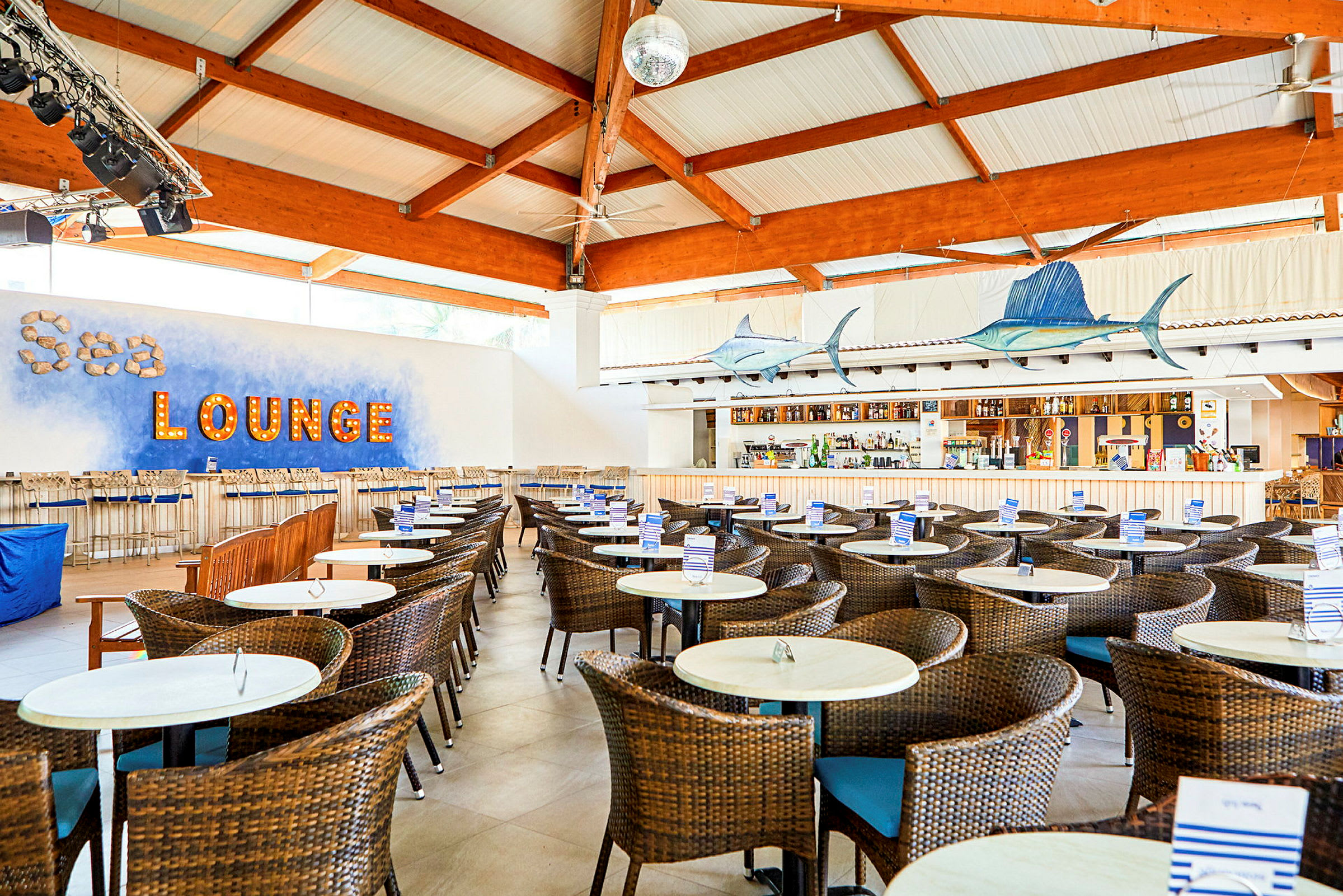 Interiör av en strandbar med rottingstolar och bord, upplyst text som lyder 'Sea lounge' på väggen. (AI-genererad text)