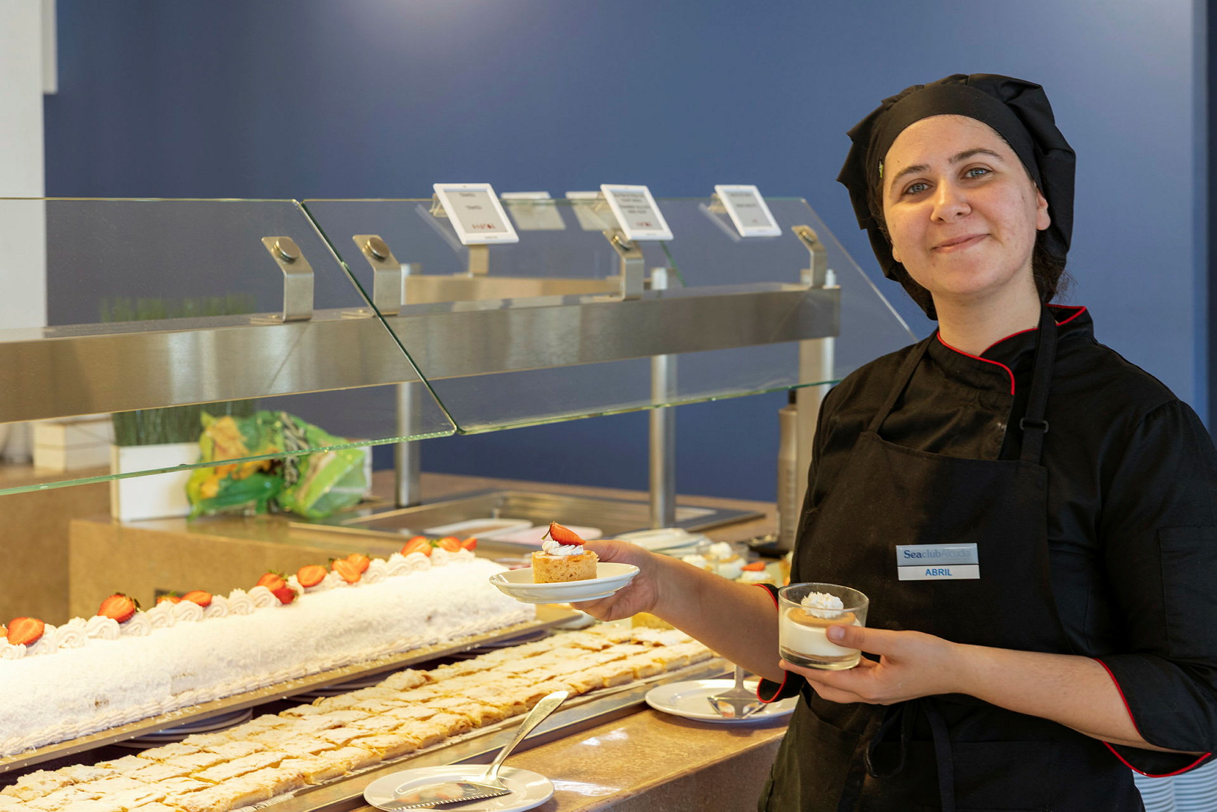 En kock klädd i svart står vid en buffé med desserter och håller i två upplagda portioner. (AI-genererad text)