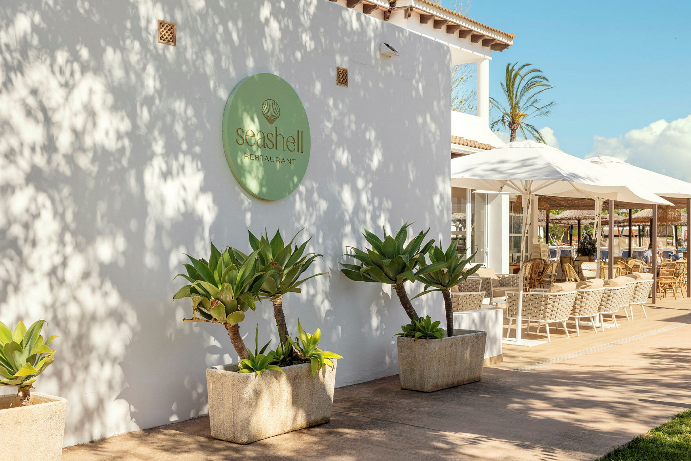 Exteriör av restaurangen Seashell med vita väggar, gröna växter och uteservering med parametriska paraplyer. (AI-genererad text)