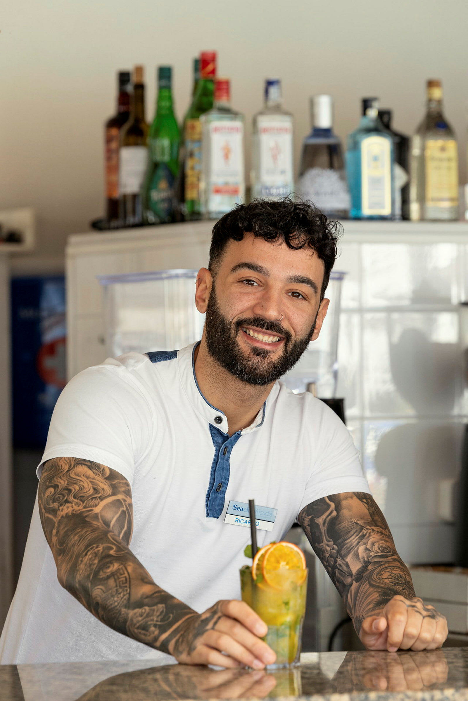 En leende man står vid en bardisk med olika spritflaskor i bakgrunden och håller i en cocktail med apelsinskiva. (AI-genererad text)