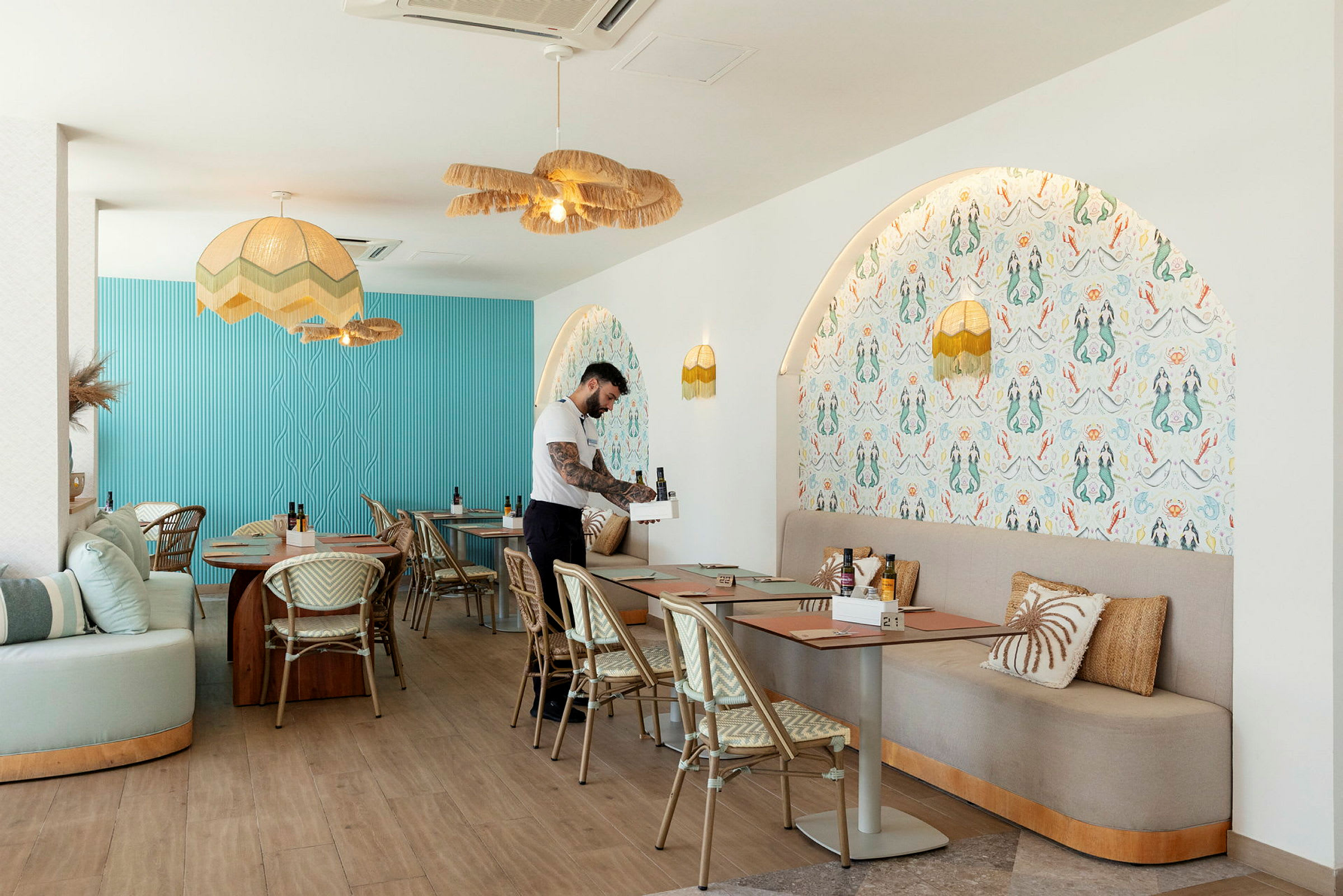 En interiör av en modern restaurang med blå och mönstrade väggar, bord med stolar och en servitör som förbereder en plats. (AI-genererad text)
