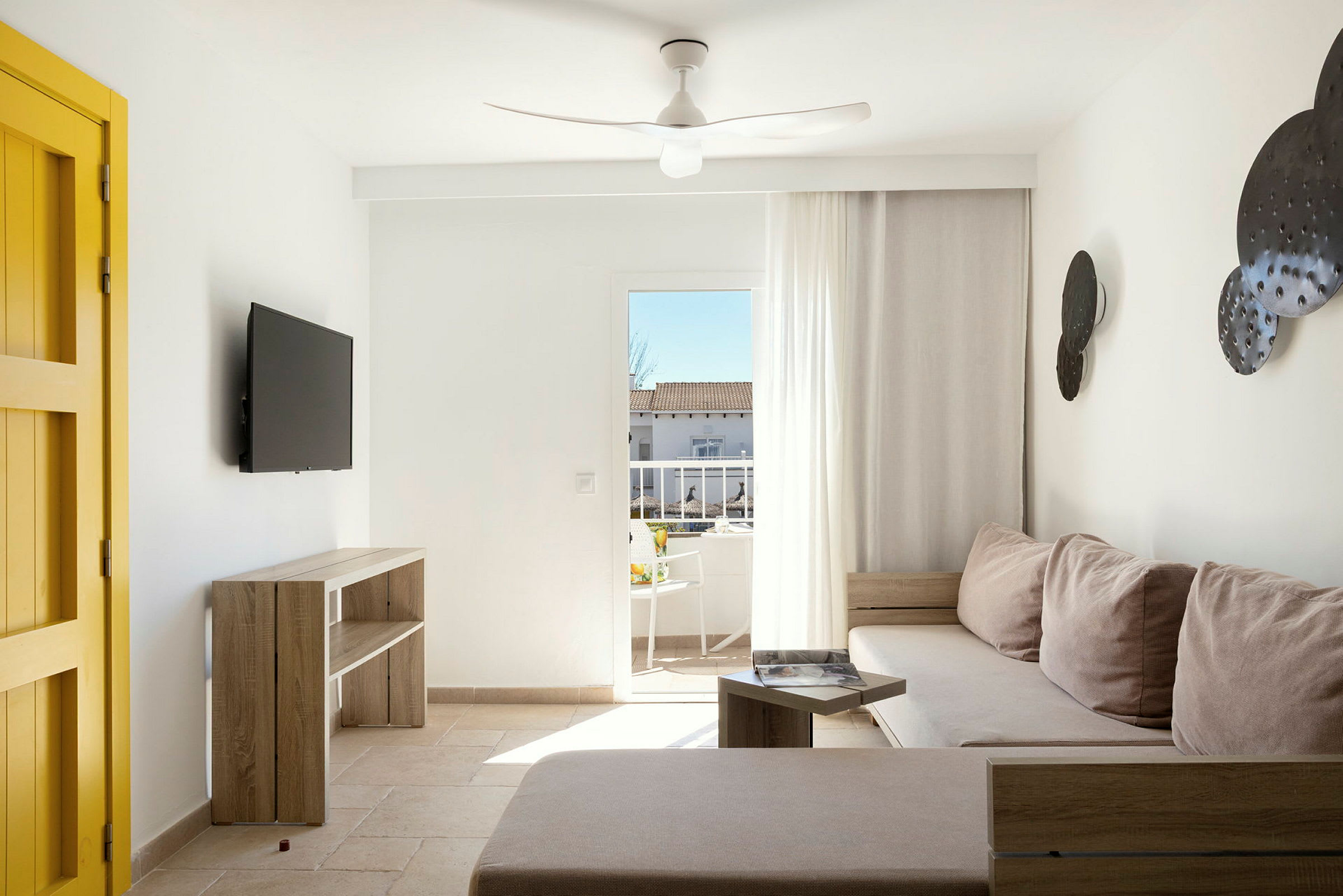 Ett ljust vardagsrum med en gul dörr, väggmonterad TV, beige soffa och direkt utgång till en balkong. (AI-genererad text)