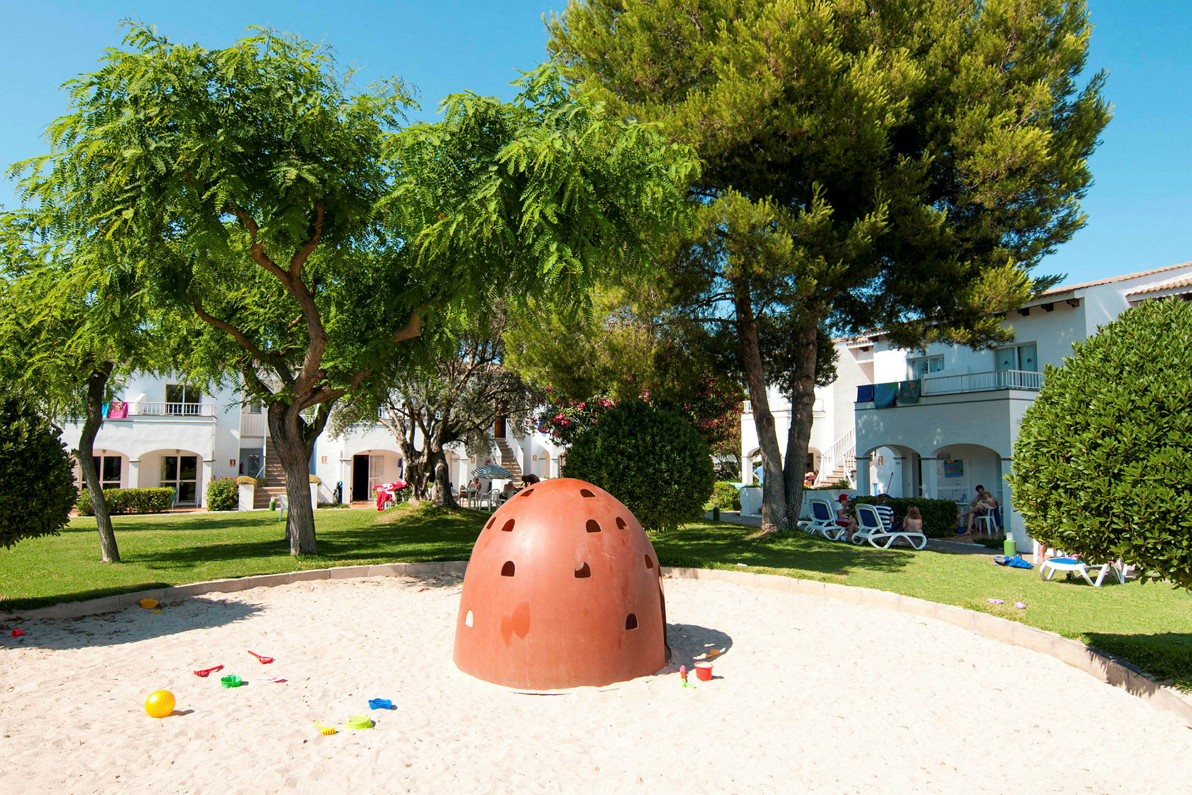 En lekplats med sand och en klätterkupol på en grön park med träd och byggnader i bakgrunden. (AI-genererad text)